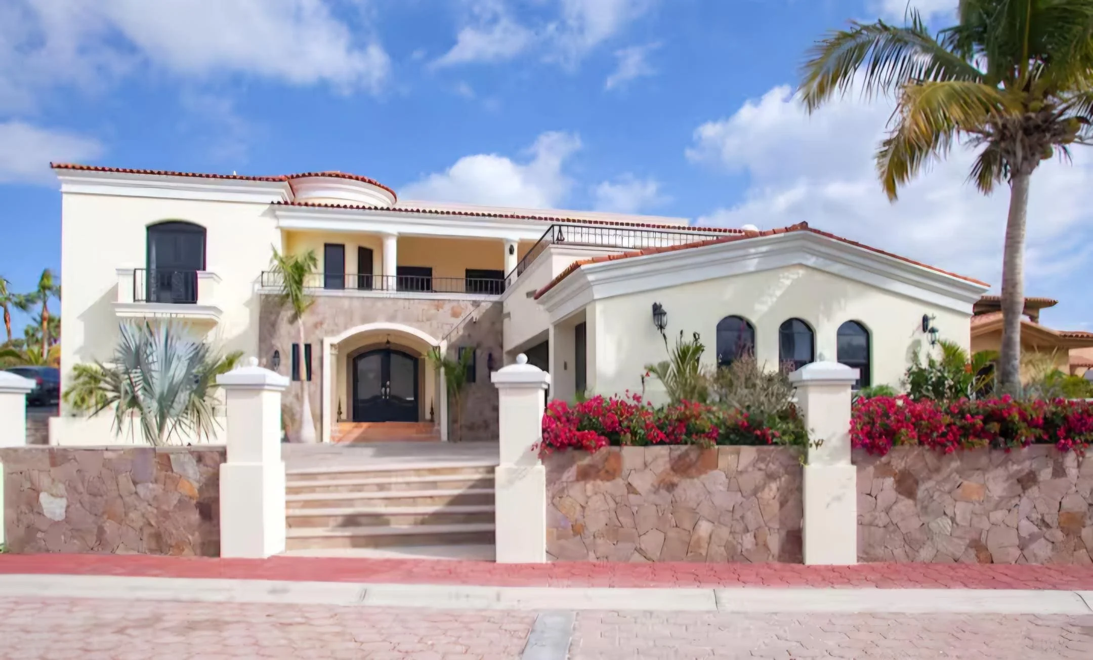 A large, luxurious, villa-style house with white walls, red tiled roof, arched windows, and a stone wall with stairs leading to the entrance. Surrounded by tropical plants and colorful flowers, with a palm tree on the right, under a partly cloudy blu