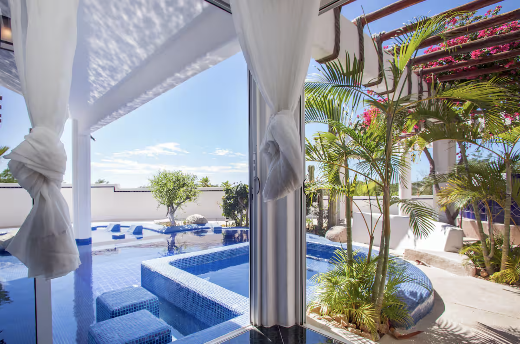 A patio with a small blue-tiled hot tub, surrounded by lush green tropical plants, with an outdoor pool in the background, under a clear blue sky.