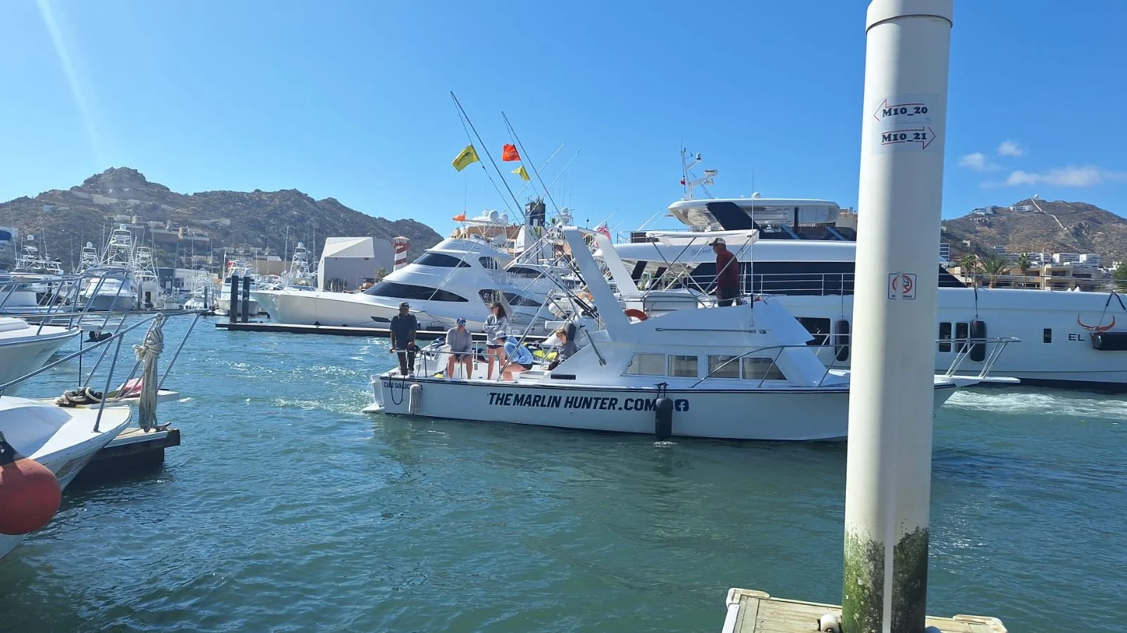Yachts docked at a marina with people on a small boat in the water against a backdrop of hills and buildings on a sunny day.