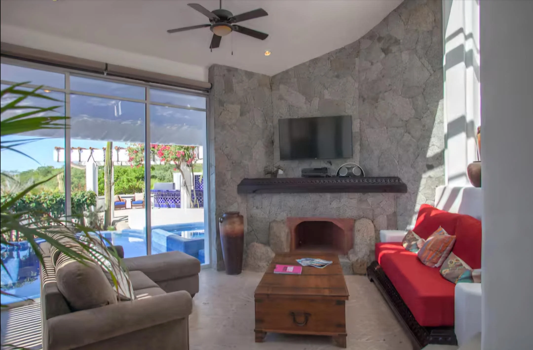 Living room with a stone fireplace, wall-mounted TV, red sofa with colorful pillows, beige armchair, wooden coffee table, large sliding glass door revealing outdoor seating and pool area, ceiling fan.