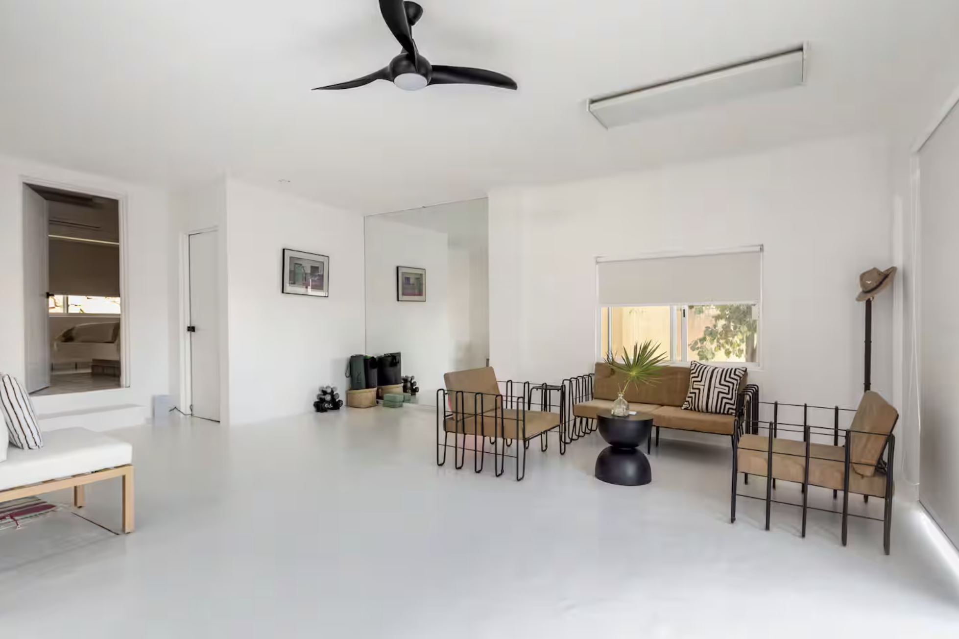 Minimalist living room with white walls and floor, black ceiling fan, and modern furniture including a beige sofa with pillows, two black metal armchairs with beige cushions, a small black round coffee table with a plant, and two framed artworks on t