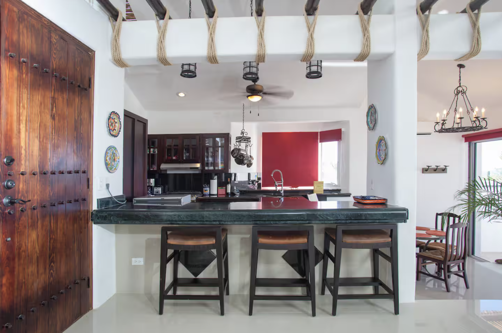 Kitchen with a dark green marble countertop, three bar stools, and wooden cabinets. Open shelving with hanging pots, ceiling fan, and a window with red blinds. Adjacent dining area with a chandelier and patio door.