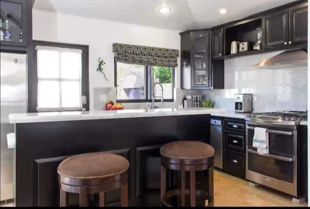 Kitchen with black cabinets, white walls, stainless steel stove, microwave, and fridge, two brown stools at a breakfast bar, window with patterned valance, and kitchen tools on countertop. The Marlin Hunter luxury villas in Cabo