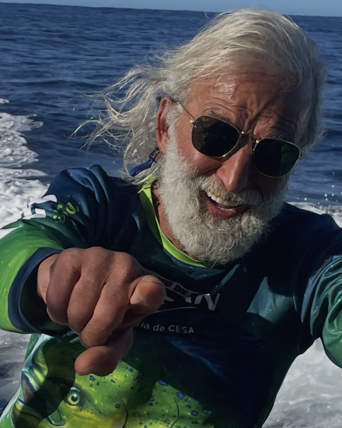 Man with white hair and beard wearing sunglasses and a colorful fishing jersey, taking a selfie on a boat with ocean in the background.