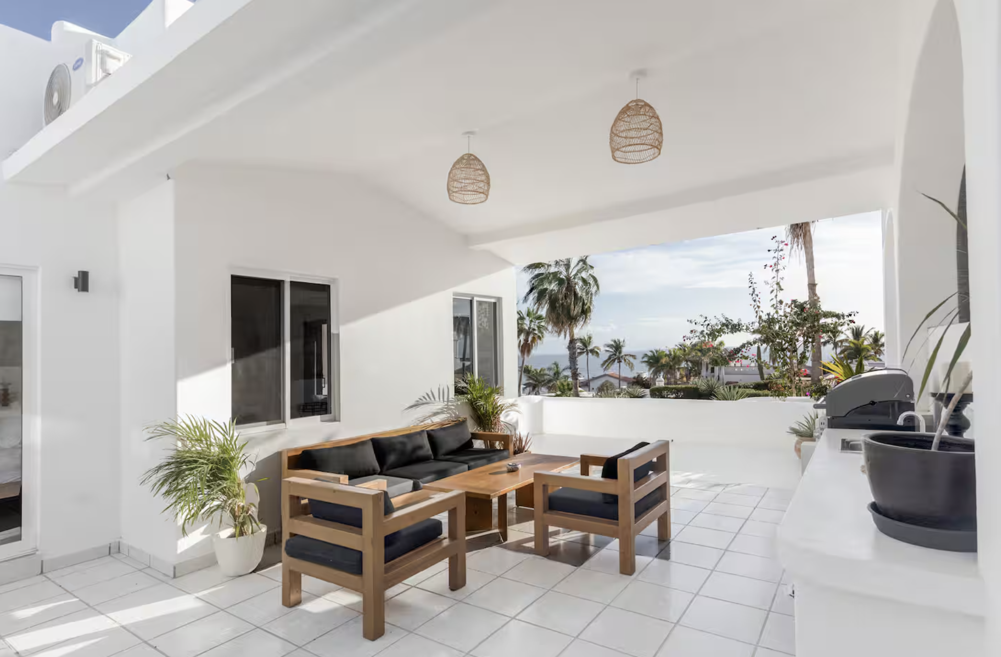 Bright outdoor patio with white walls and tile floor, outdoor seating with wooden furniture and black cushions, potted plants, barbecue grill, and a view of palm trees and the ocean in the background.