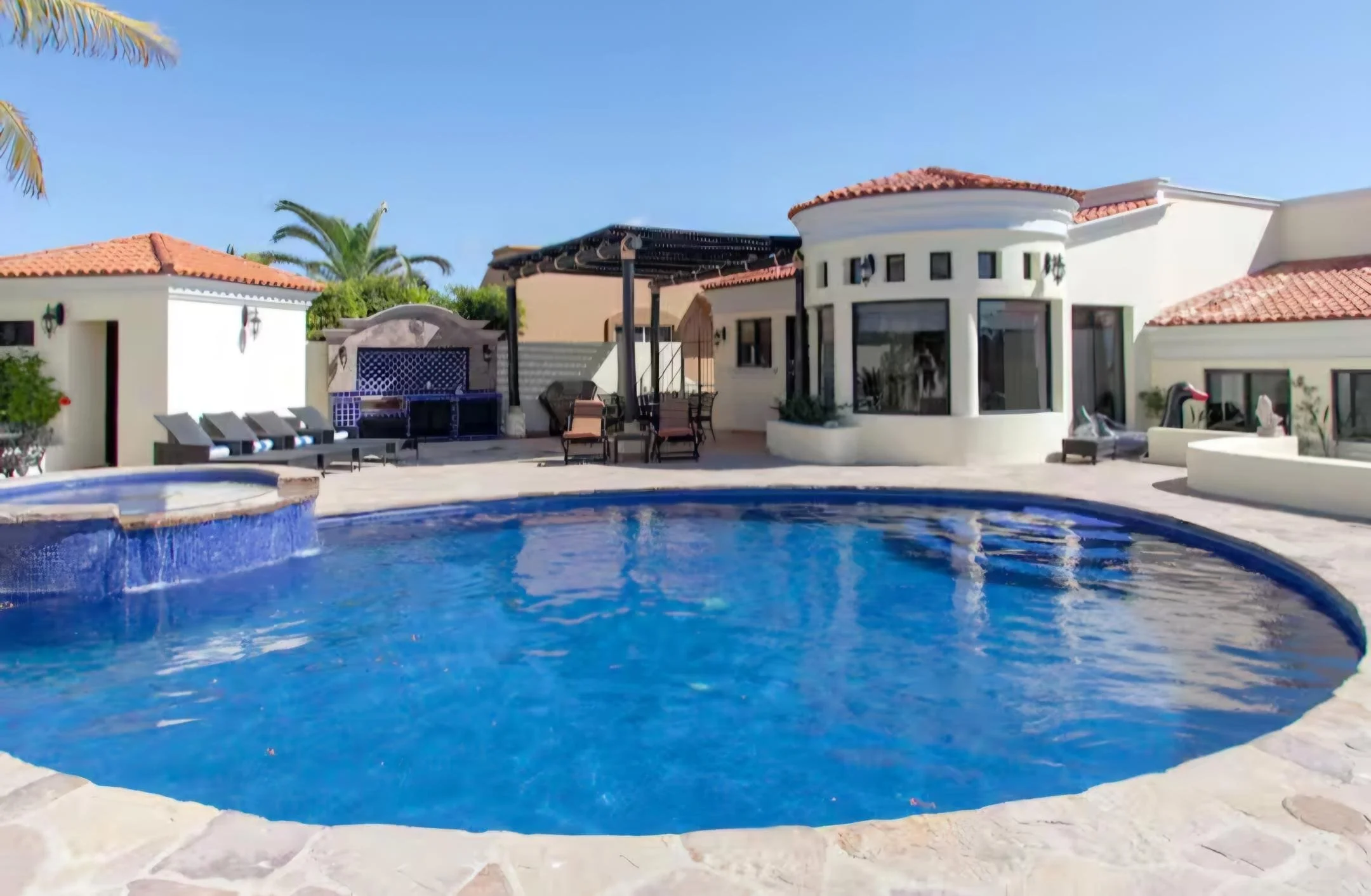 Luxury mansion with a swimming pool in the backyard, surrounded by white walls, lounge chairs, and palm trees, under a clear blue sky. The Marlin Hunter luxury villas
