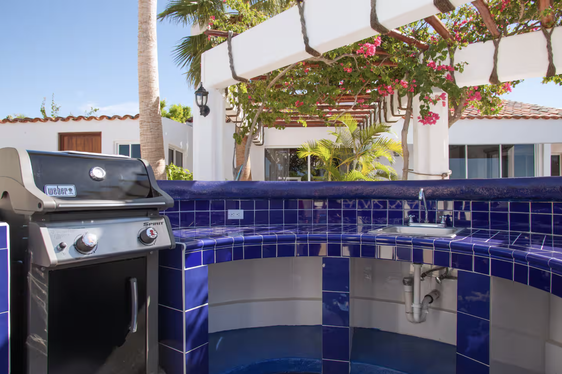 Outdoor kitchen with a propane grill and blue tiled countertop, surrounded by palm trees, flowering vines, and a white building with a red tile roof.