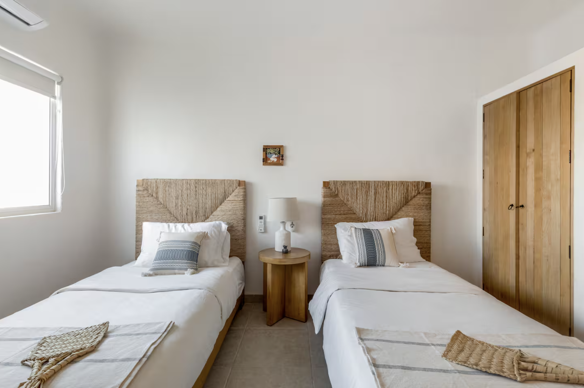 A bedroom with two single beds with woven headboards, a small round wooden nightstand with a lamp between the beds, a small framed artwork above the nightstand, a window with a white blind on the left, and a closed wooden closet door on the right. Th