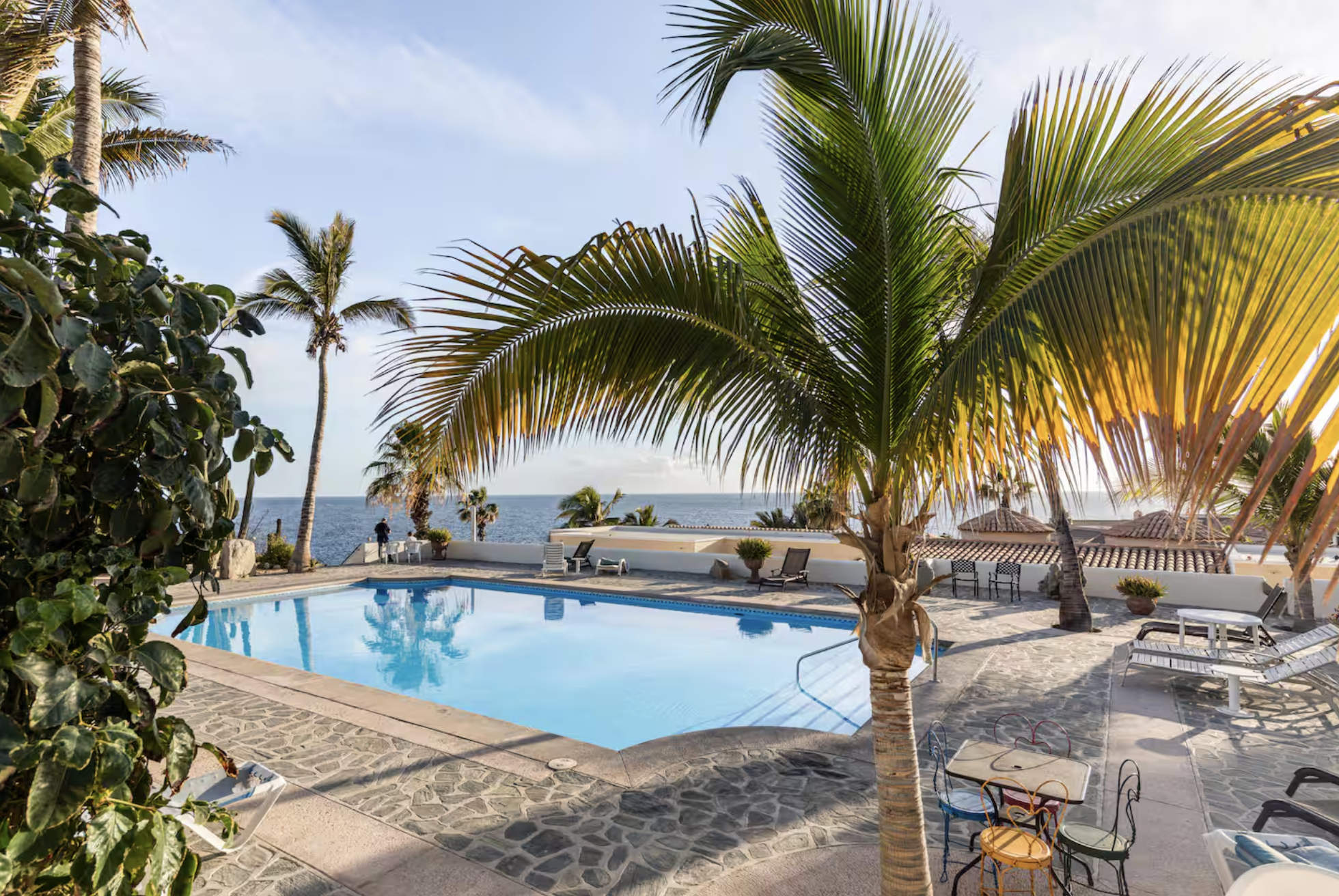 Poolside area with lounge chairs, tables, palm trees, and a view of the ocean in the background.