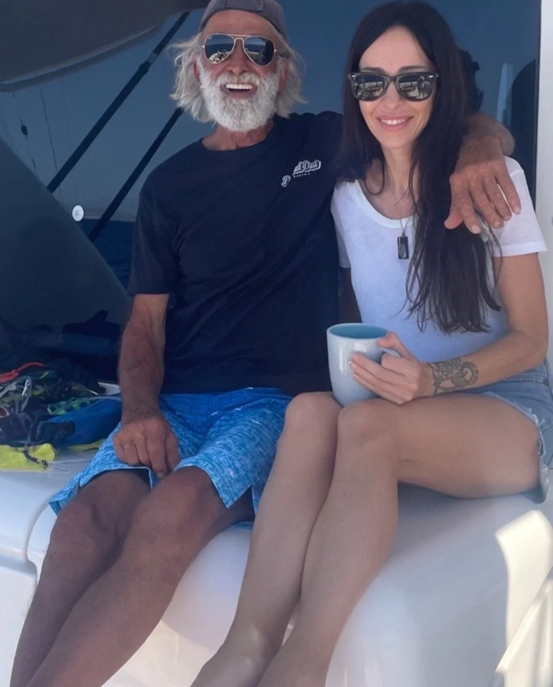 A smiling older man with a white beard wearing sunglasses and a backwards cap, sitting next to a smiling young woman with long dark hair, wearing sunglasses and holding a mug, on a boat with a blue sky in the background.