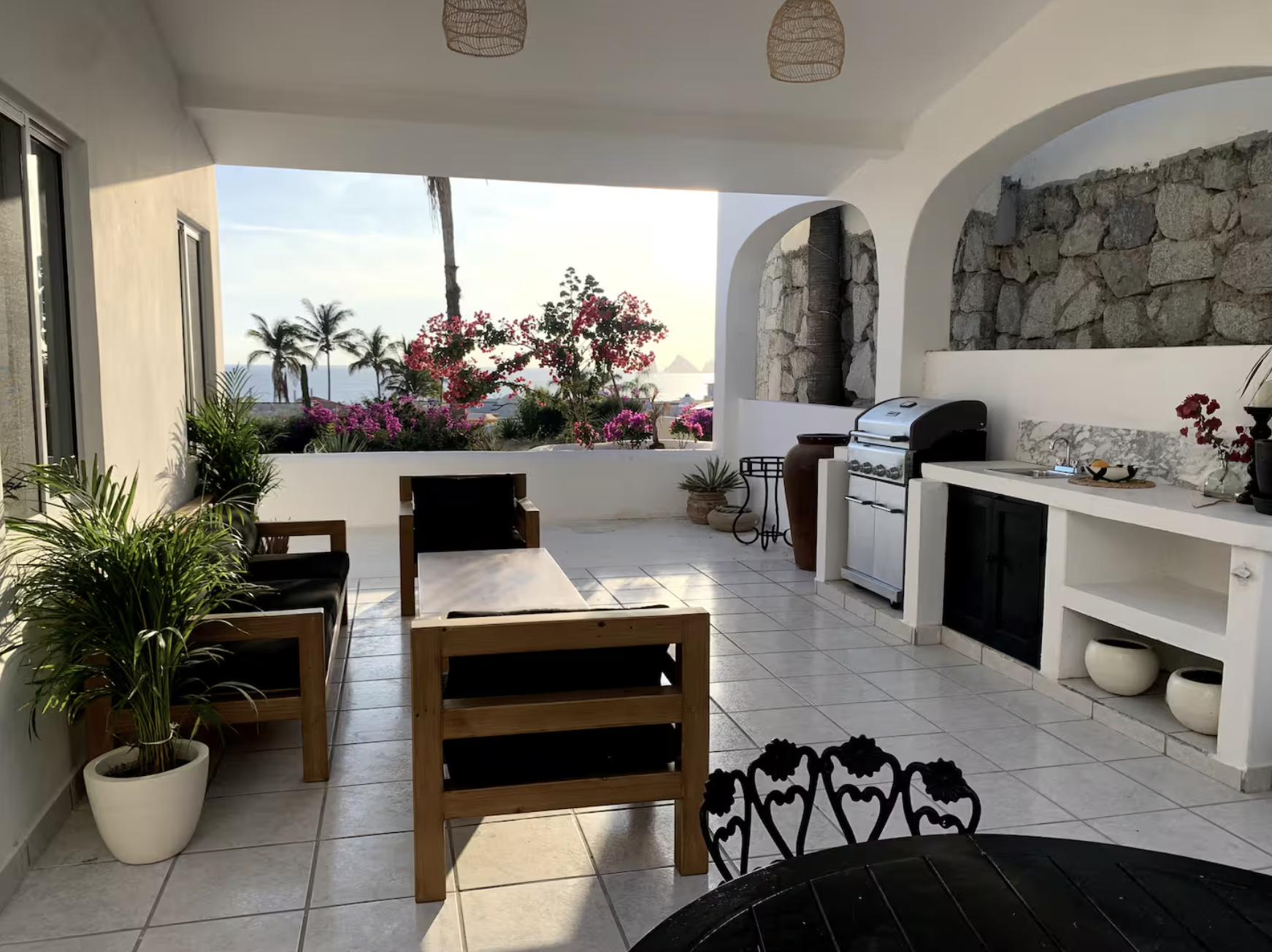 Covered outdoor patio with wooden and metal chairs, a table, potted plants, a barbecue grill, and view of palm trees and ocean in the background.