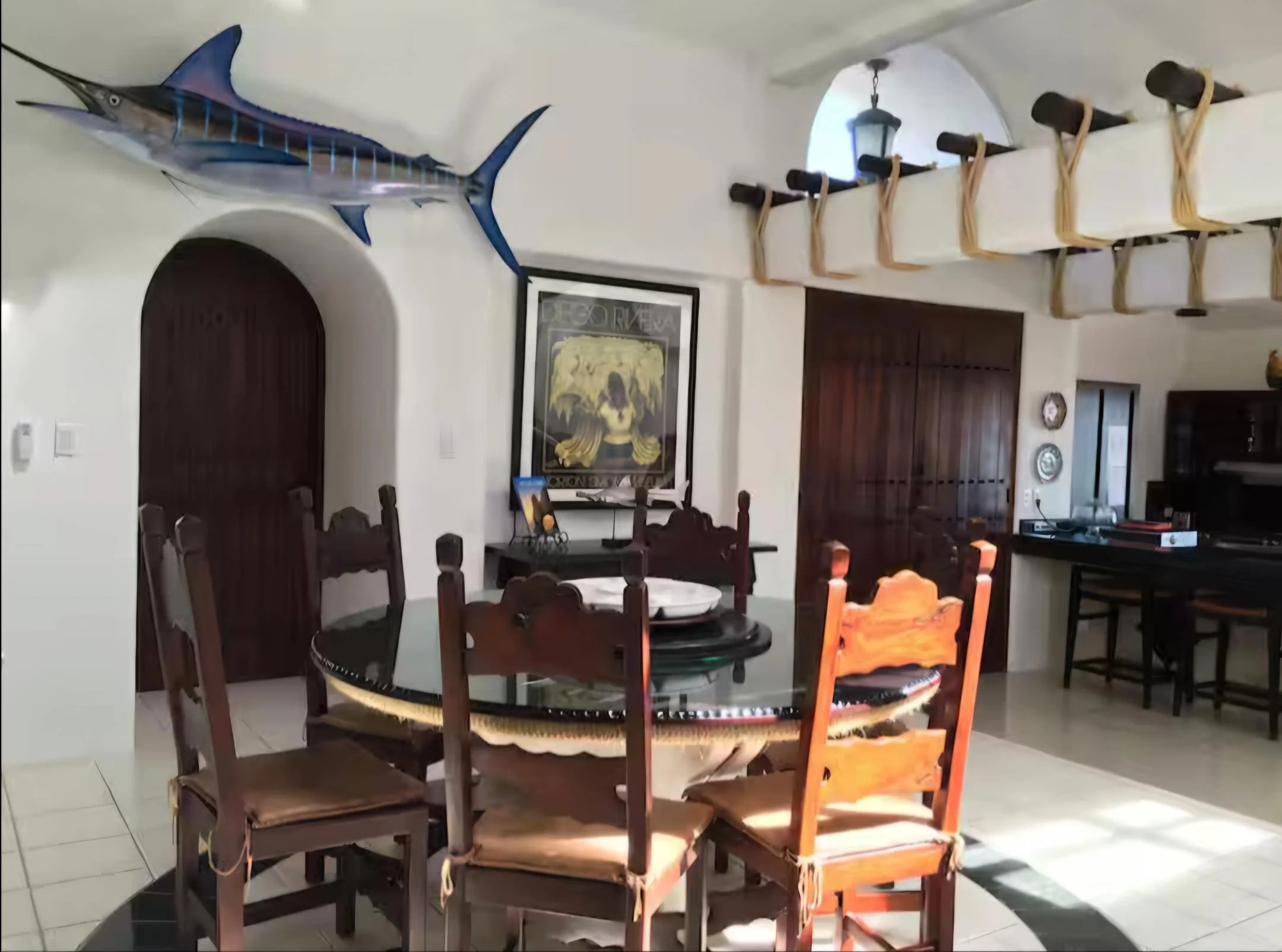 A dining room with a round glass-top table surrounded by six wooden chairs with cushioned seats. A framed artwork hangs on the wall, and a mounted marlin fish is displayed above an arched doorway. The room features a ceiling with a wooden beam and ro