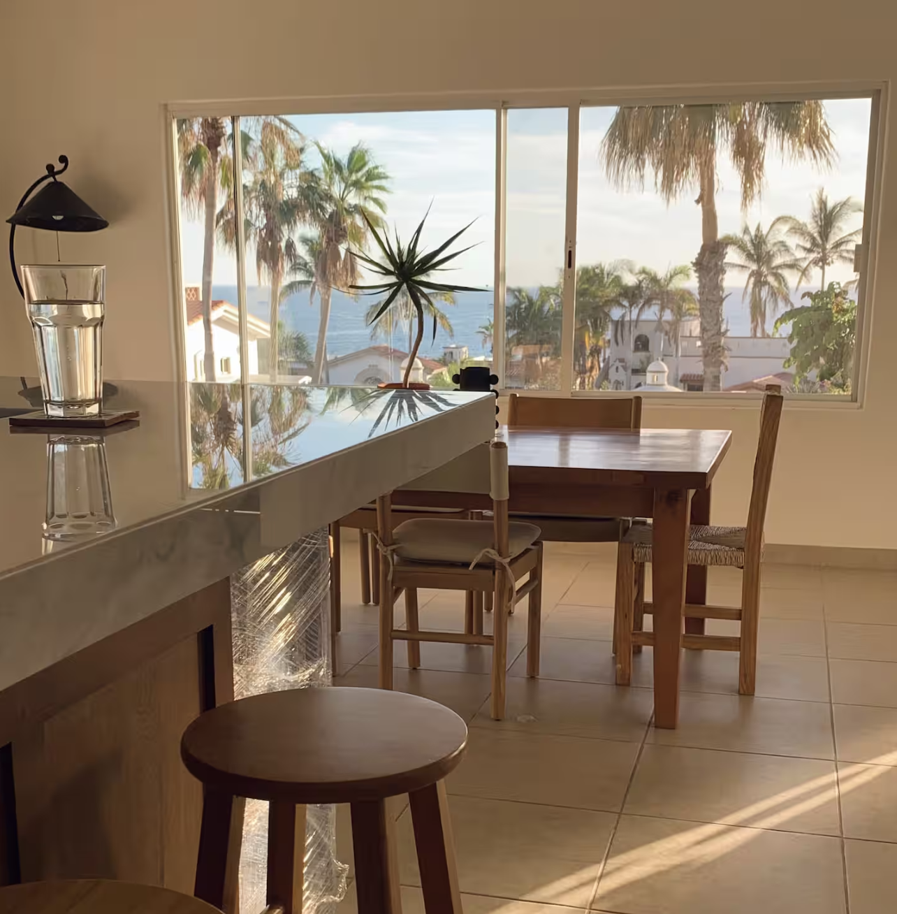 Interior of a room with a large window showing palm trees and a view of the ocean, wooden table and chairs, a bar or counter with a glass of water, and a potted plant.