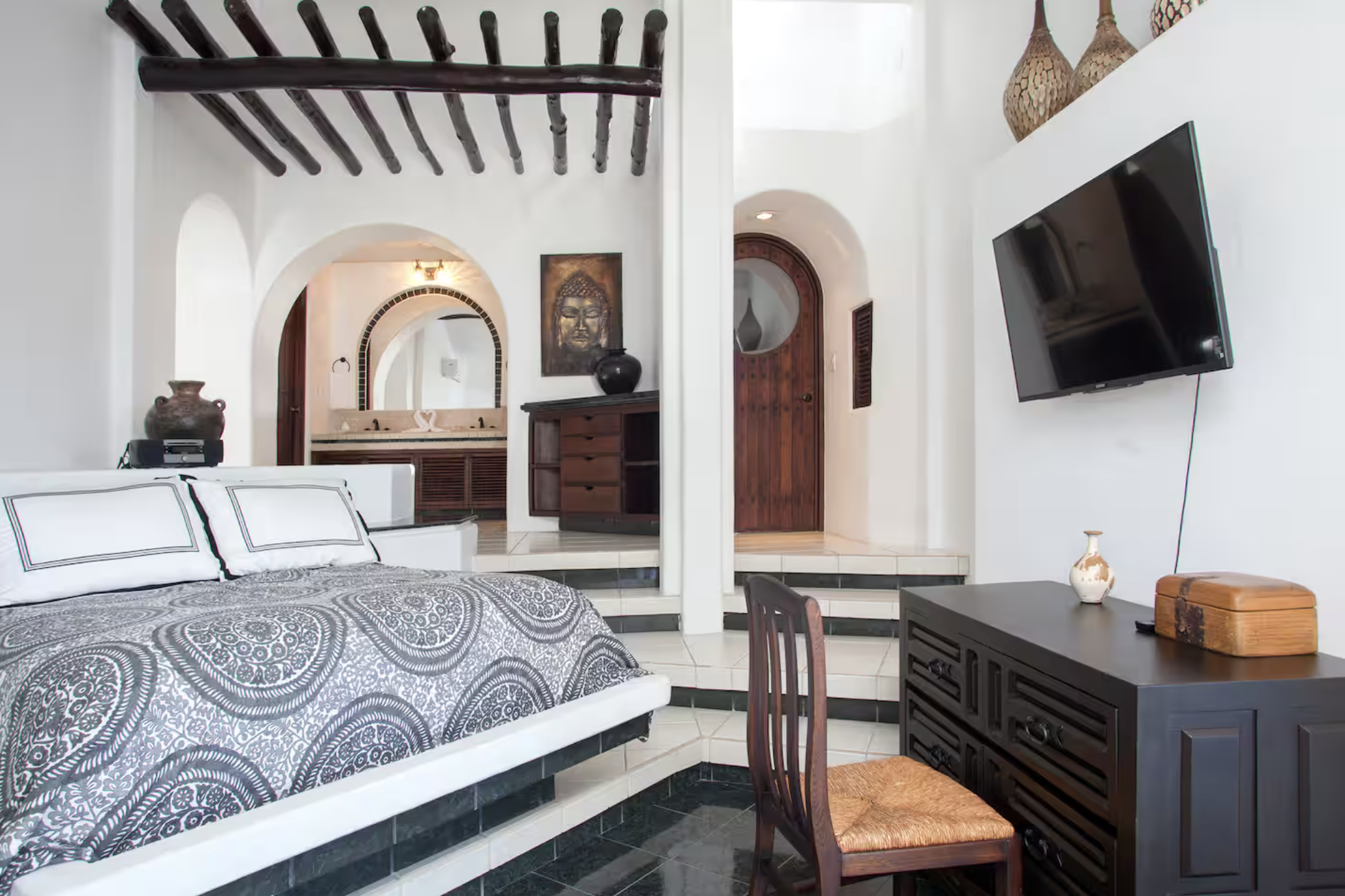 A bedroom with a bed, wooden furniture, a flat-screen TV on the wall, decorative vases, and an arched doorway leading to a bathroom with a sink and mirror.