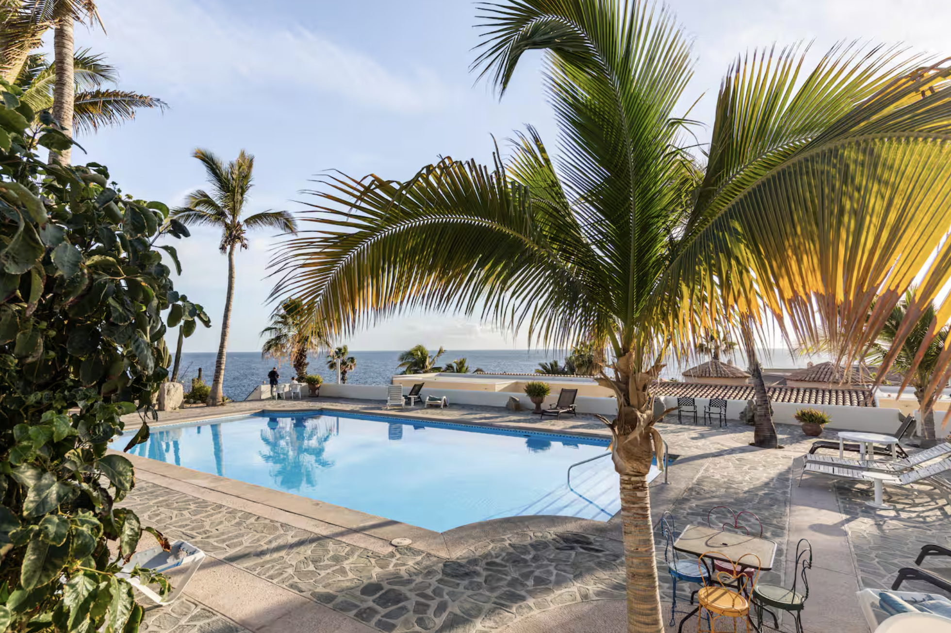 A swimming pool surrounded by palm trees and outdoor furniture, with the ocean in the background under a partly cloudy sky.