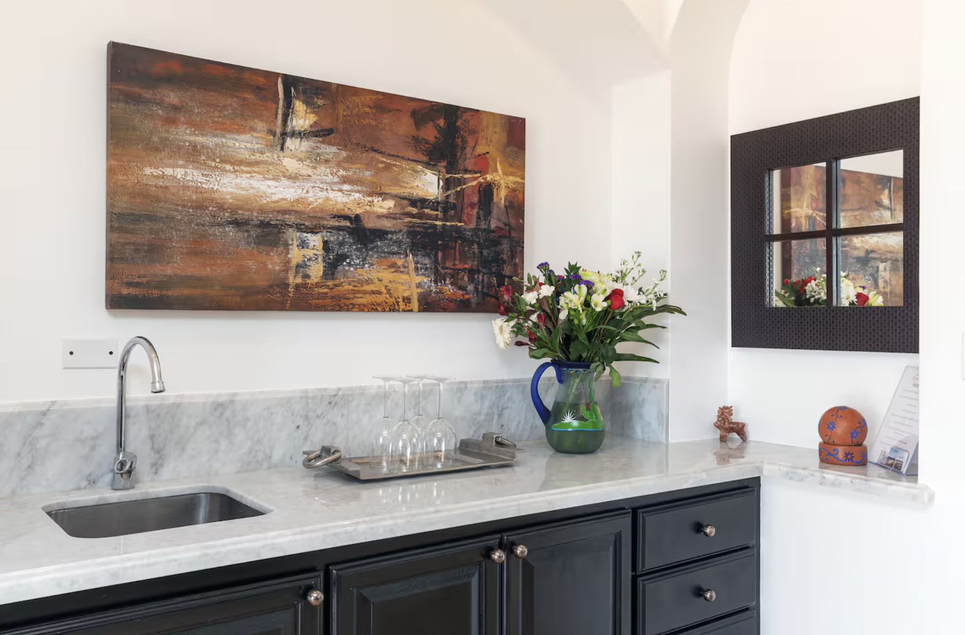 A kitchen countertop with a marble surface, a sink with a chrome faucet, and a drawer cabinet below. On the counter, there is a tray with upside-down wine glasses and a colorful floral bouquet in a blue vase. Behind the bouquet, the wall features a l