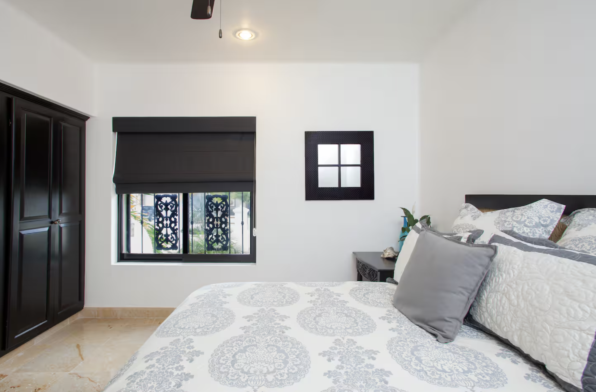 A bedroom with white walls, a black wardrobe on the left, a window with black decorative wrought iron bars and a black window shade, a small black nightstand with a seashell, a plant, and a blue spray bottle on it, a bed with white and gray floral be