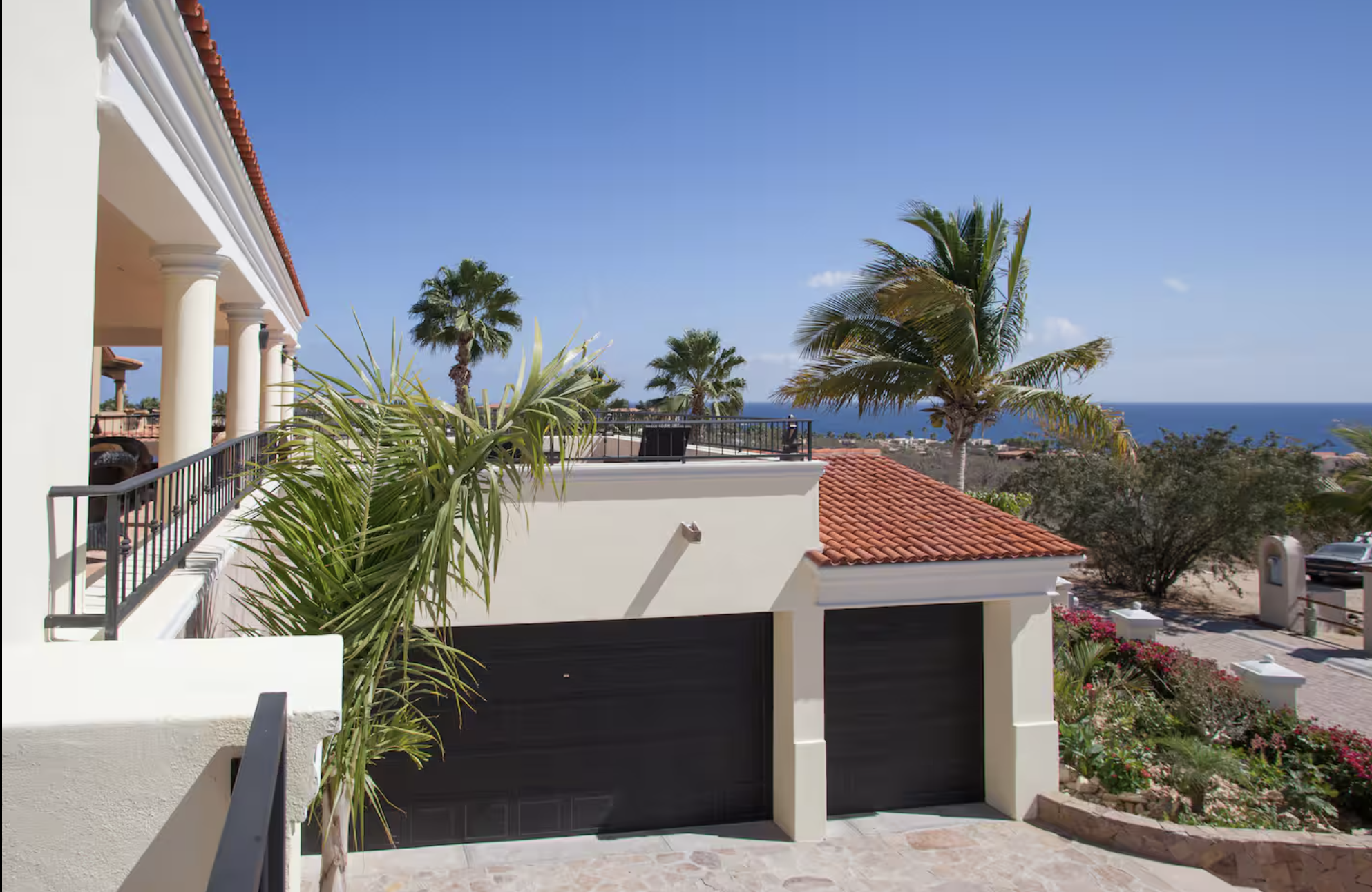 View of a Mediterranean-style house with white exterior, red-tiled roof, and black garage doors, surrounded by palm trees and lush landscaping, with the ocean in the background under a clear blue sky.