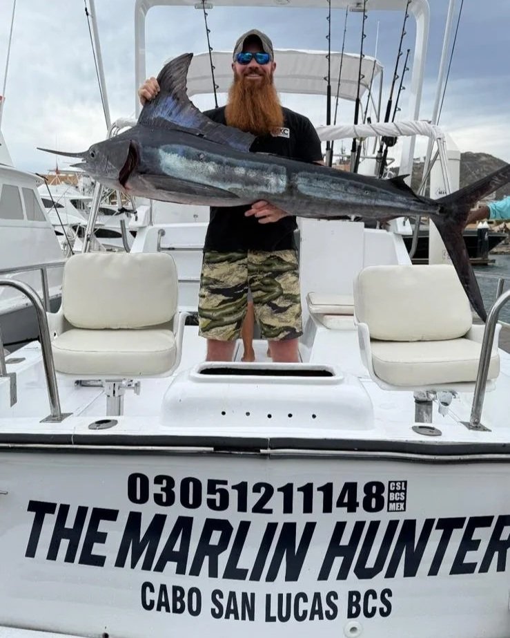 Man with a long beard, sunglasses, and dark T-shirt holding a large fish on a boat in Cabo San Lucas.