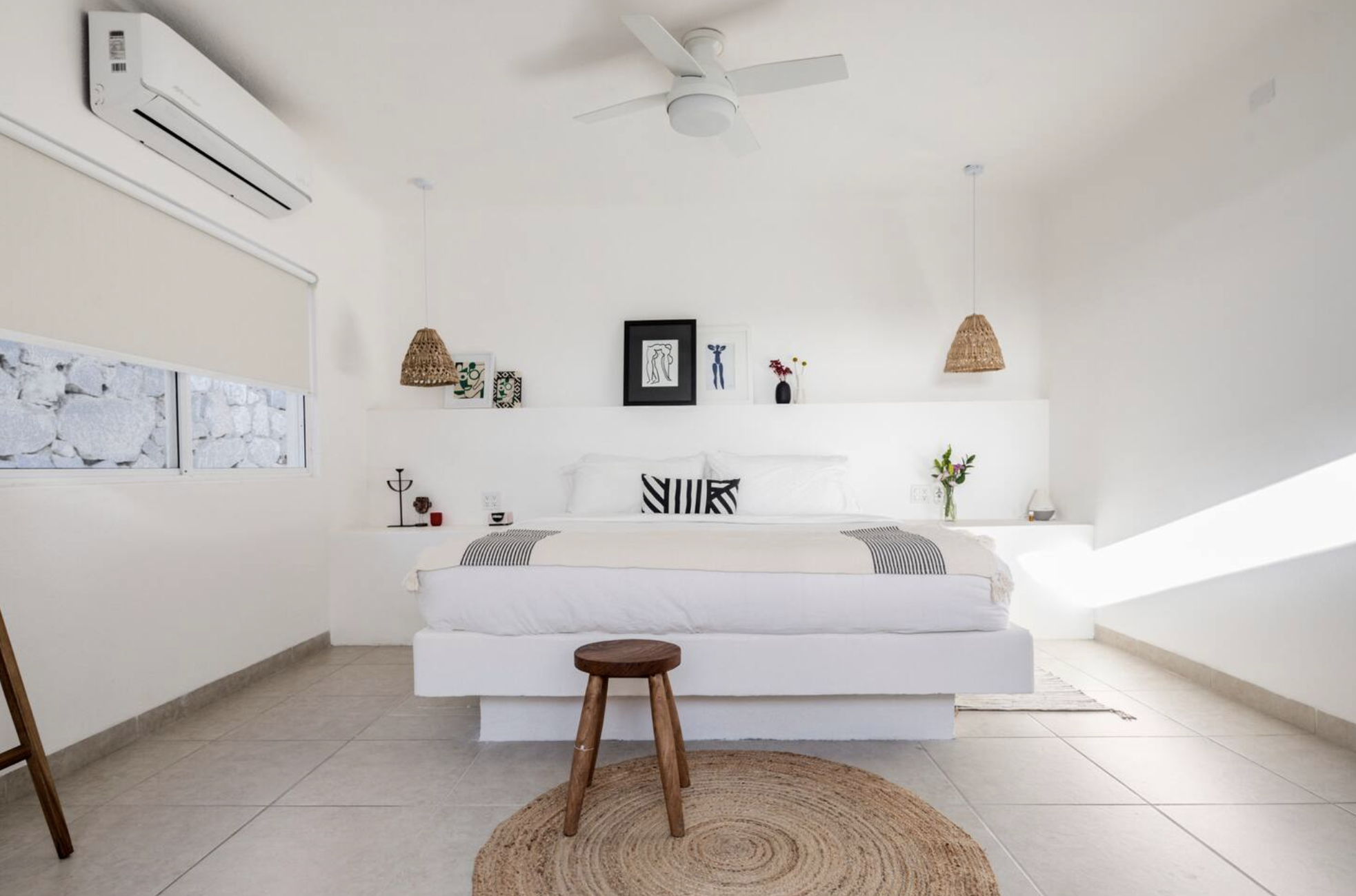 Minimalist bedroom with white walls, bed with black and white striped pillow, wooden stool, round woven rug, artwork, potted plants, air conditioner, ceiling fan, and window with a blind.
