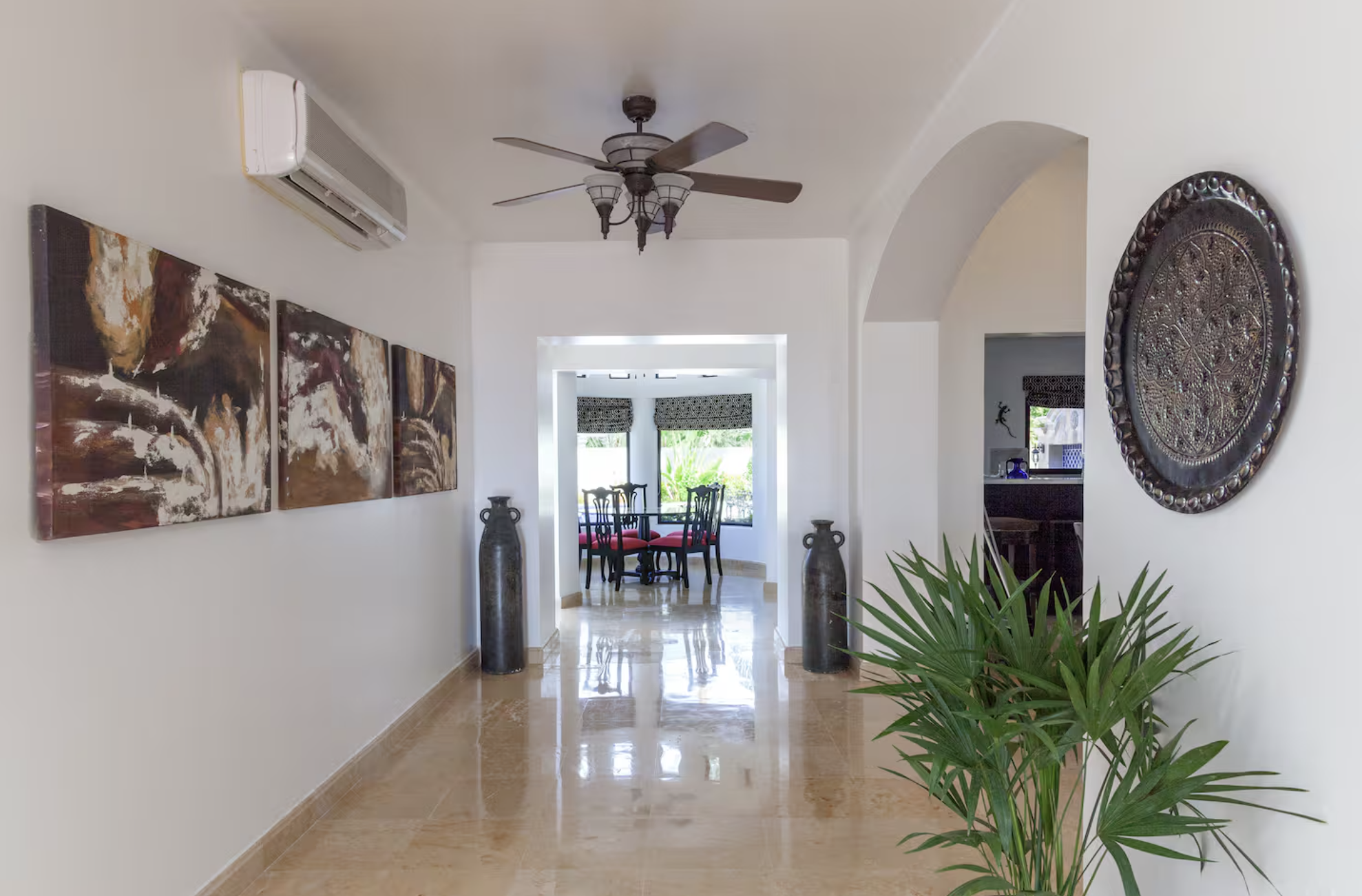 A hallway leading to a dining area with a round table and four chairs. The hallway has cream-colored walls, a ceiling fan, artwork on the left, a large metal decorative plate on the right wall, large vases on either side of the hallway entrance, and 