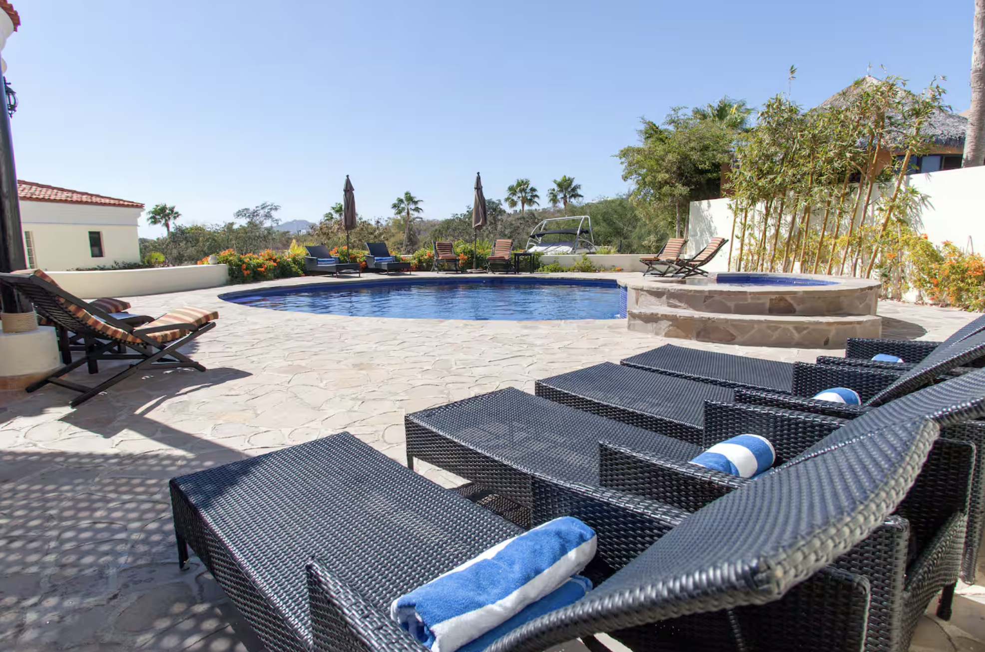 Poolside area with several black wicker lounge chairs with blue and white towels, a hot tub with stone surround, a round swimming pool, umbrellas, and tropical plants under a clear sky.