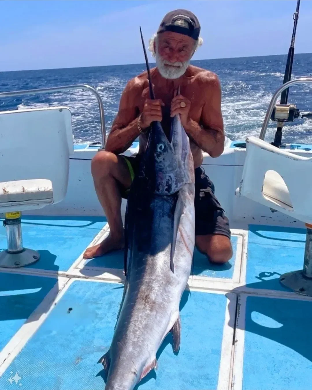 An elderly man with white hair and beard, wearing a backwards cap, kneeling on a boat deck, holding a large fish he caught while fishing in the open ocean on a sunny day.