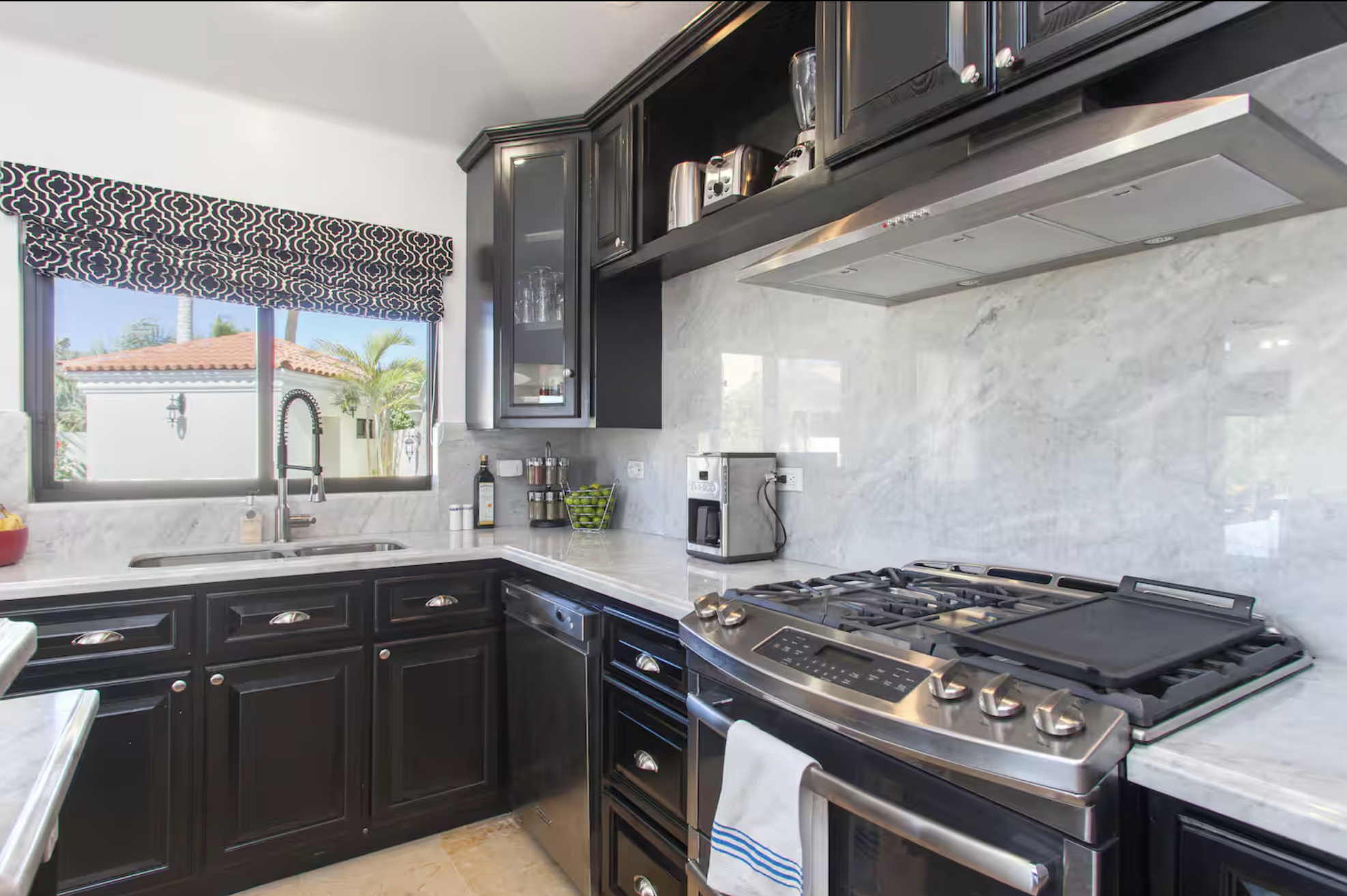 Modern kitchen with black cabinets, stainless steel stove, marble backsplash, window with patterned valance, and countertop with small appliances and decor.