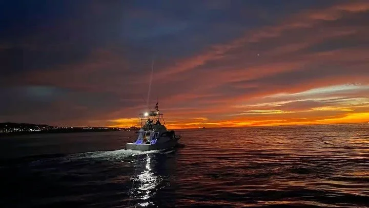 A boat sailing on the ocean during sunset with vibrant orange and purple sky and a distant shoreline on the horizon. The Marlin Hunter fishing charters