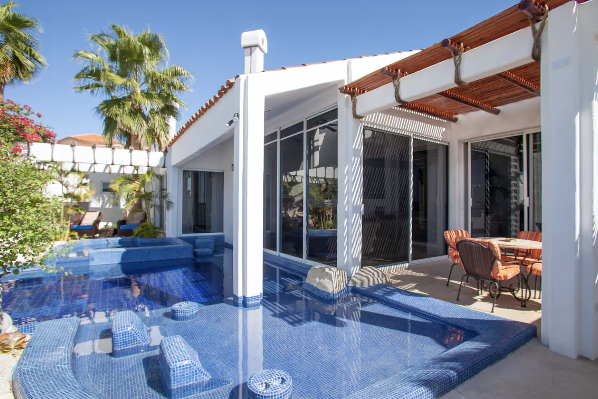 A backyard with a swimming pool, outdoor seating area, and lush greenery under a clear blue sky.
