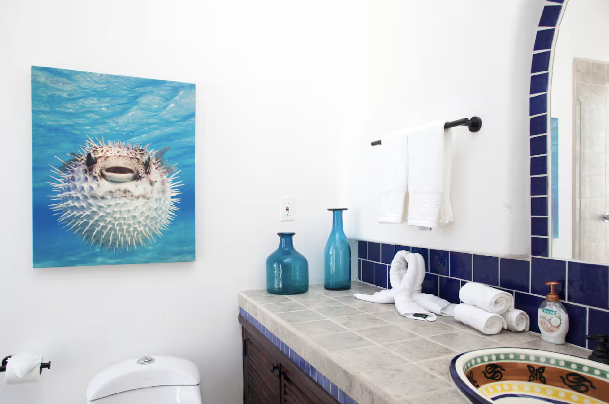 A bathroom with a pufferfish artwork on the wall, a countertop with blue vases, rolled towels, a sink with decorative design, and a soap dispenser.