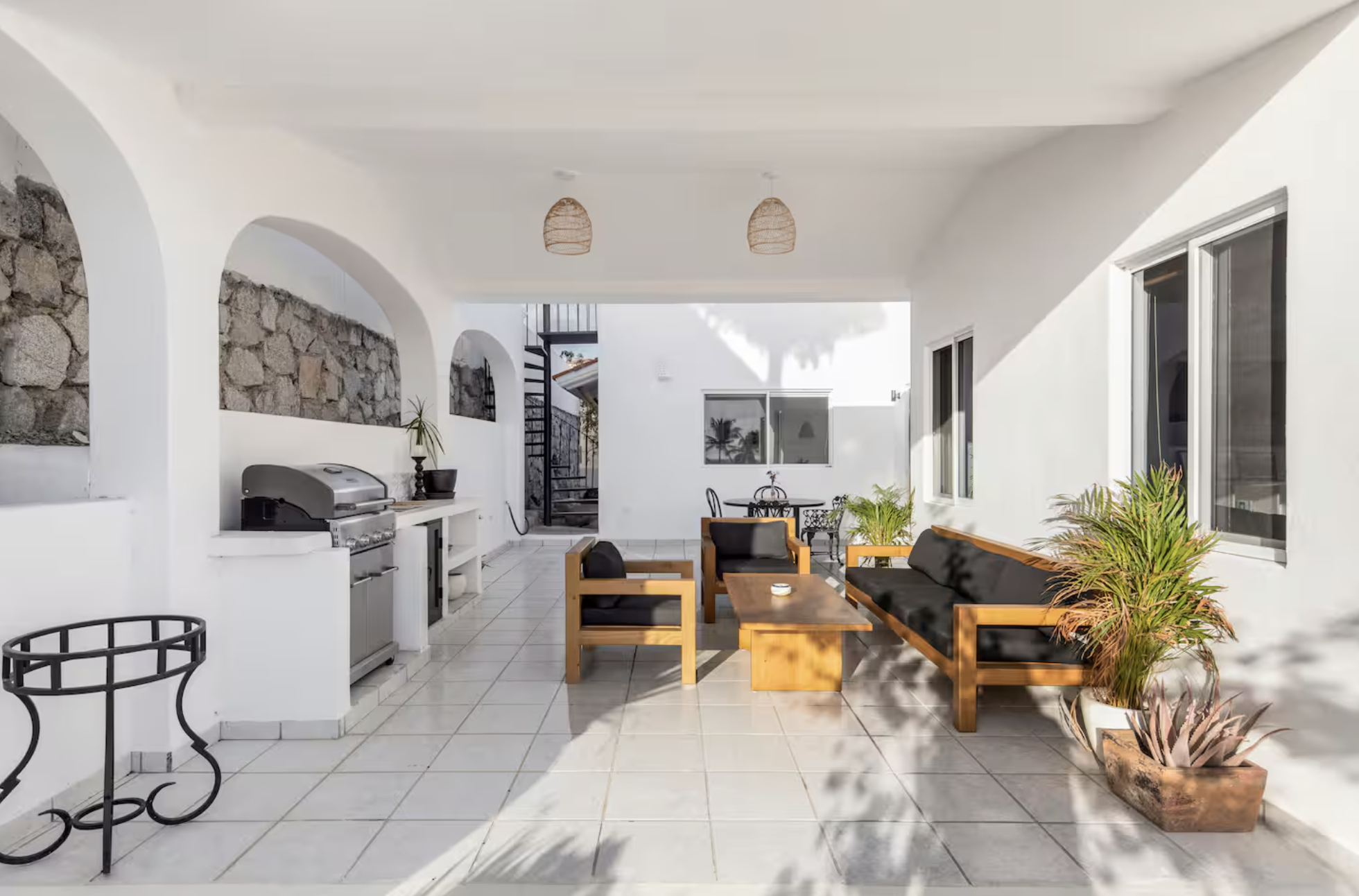 Outdoor patio with white walls, stone accents, wooden furniture with black cushions, potted plants, and a barbecue grill.