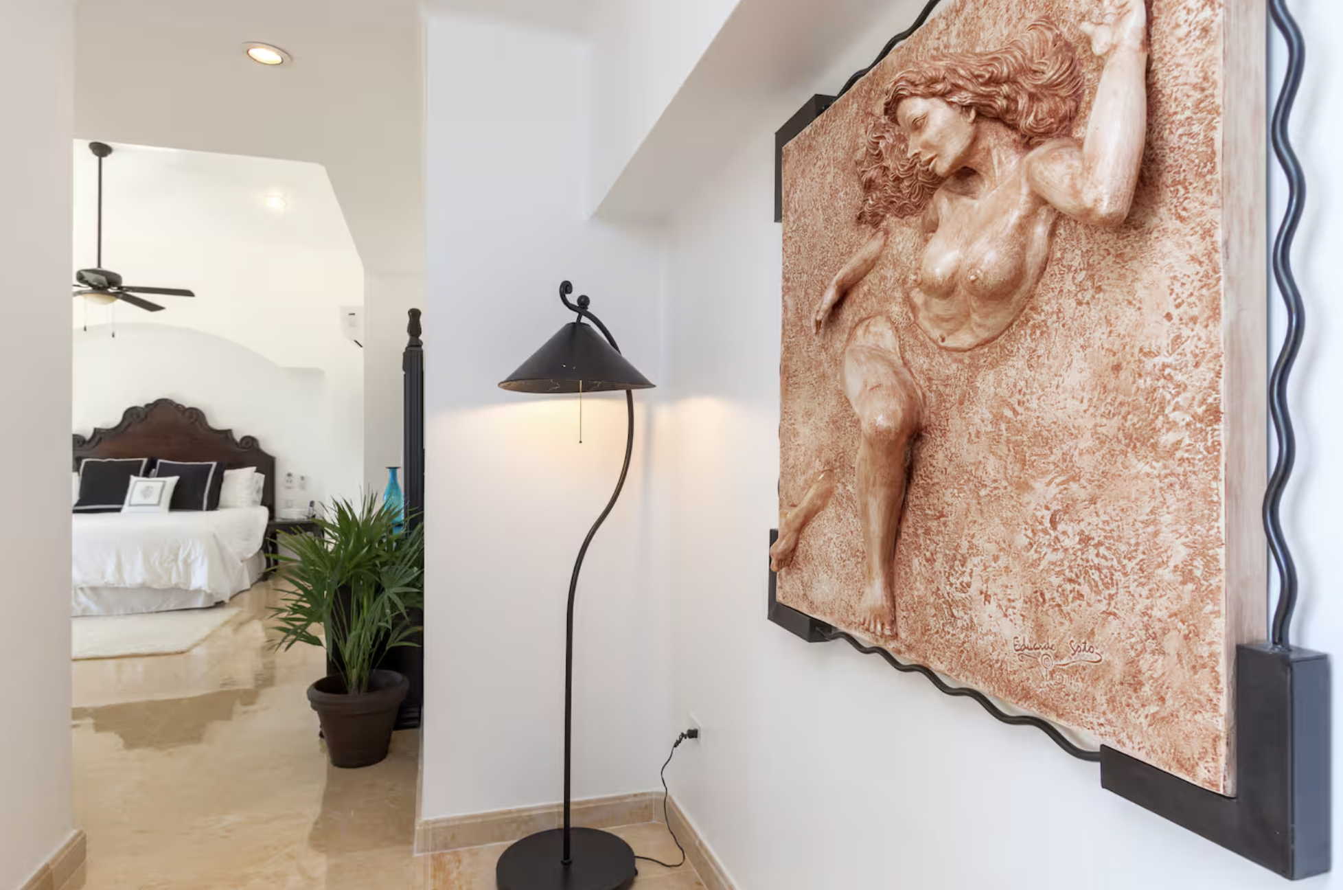 Interior view of a bedroom with a bed, drawing a large rustic wall sculpture of a woman with flowing hair, a lamp, and a potted plant.