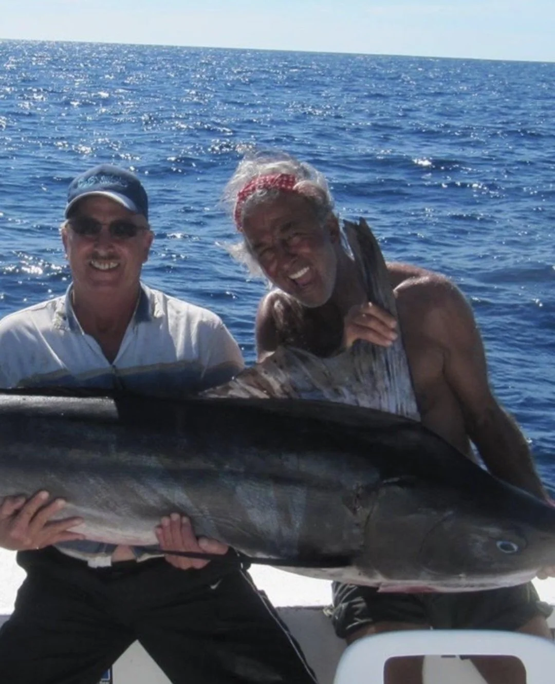 Two men smiling and holding a large fish on a boat in the ocean. The Marlin Hunter fishing charter