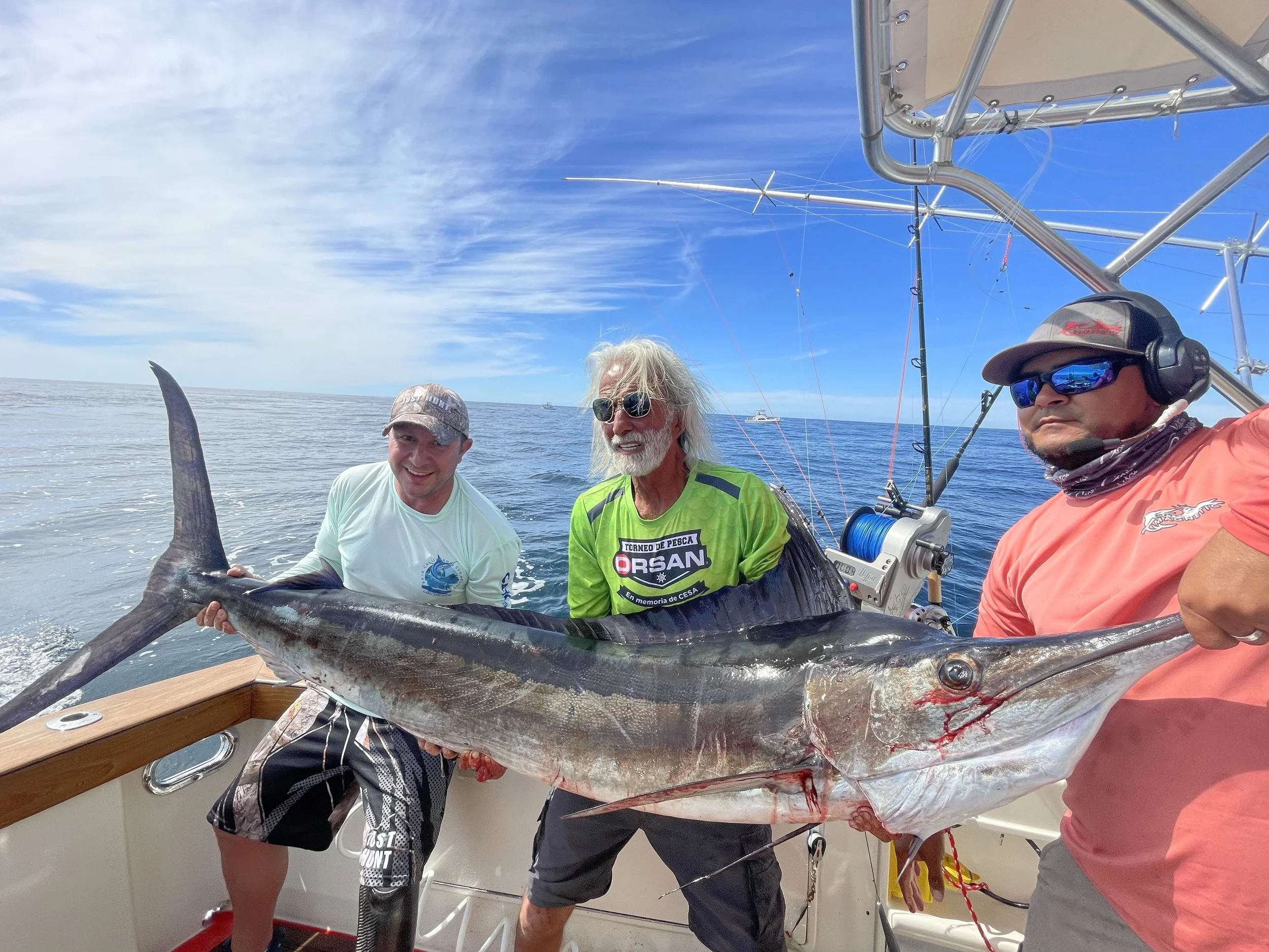 Three fishermen on a boat holding a large swordfish with an ocean and blue sky background.