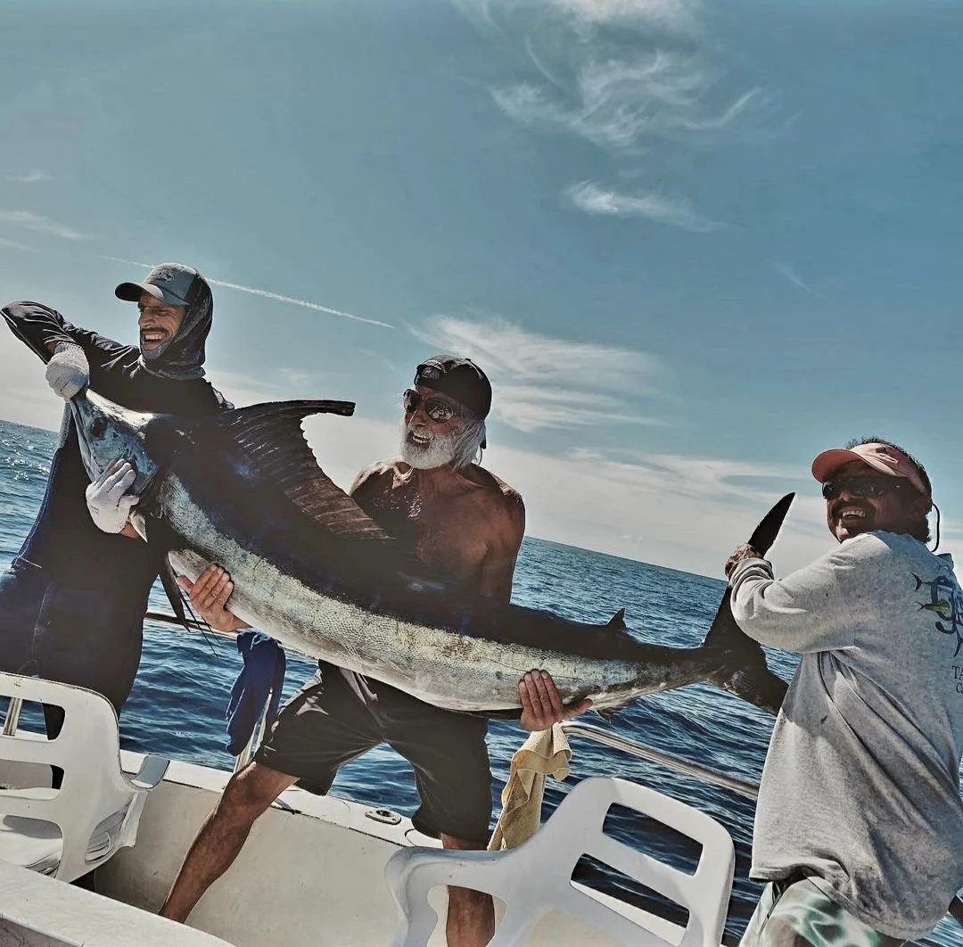 Three men on a boat holding a large fish, with the ocean and partly cloudy sky in the background. | The Marlin Hunter Fishing Charter Day Trip from Cabo