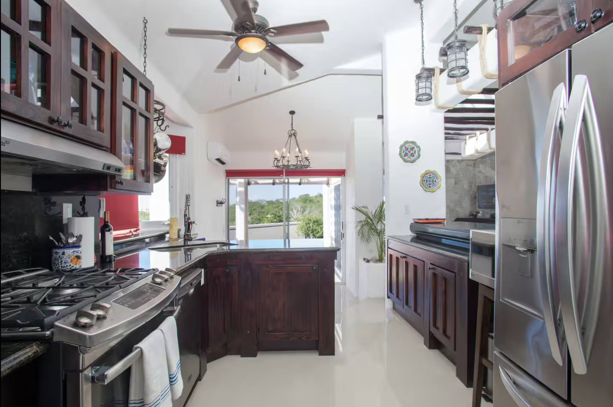 Modern kitchen with dark wood cabinets, stainless steel appliances, and bright decor. There is a ceiling fan, hanging lanterns, and a view of a balcony with greenery.