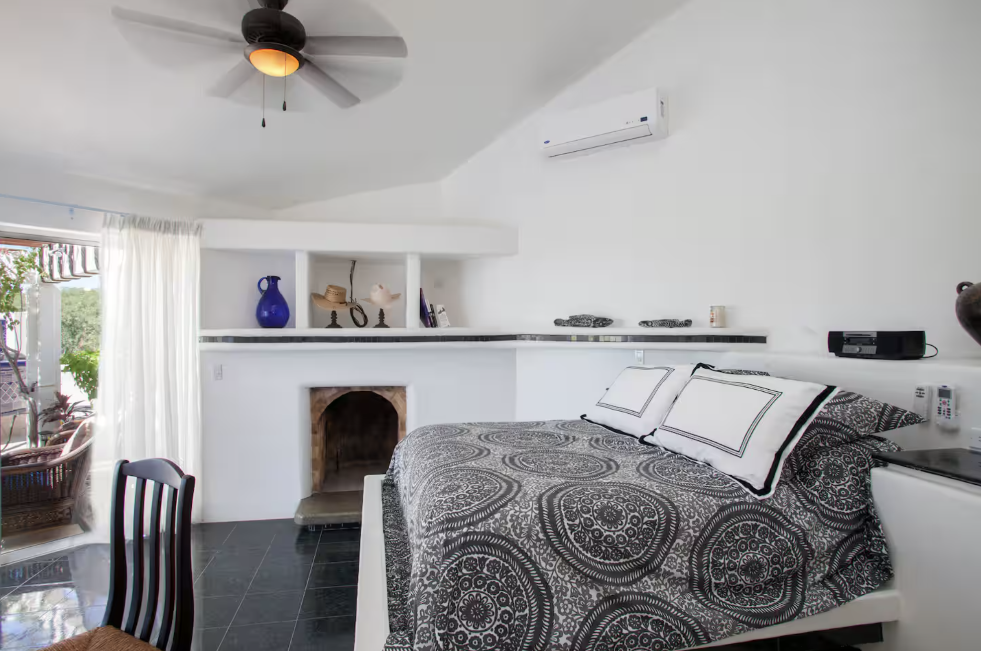 A bedroom with a white bedspread featuring black and white patterns, pillows, a fireplace, a ceiling fan with dark blades, and an air conditioner mounted on the wall. There are white curtains partially covering a sliding glass door leading outside.