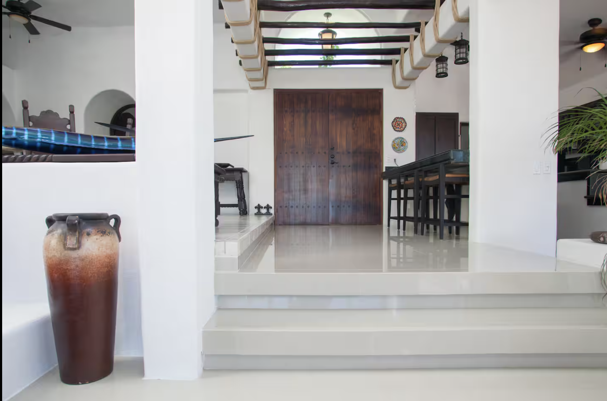 Interior view of a modern home with a wooden front door, white walls, and a tiled floor. Features include a large ceramic vase, a kitchen table with chairs, decorative plates on the wall, and ceiling fans and lantern-style lights.
