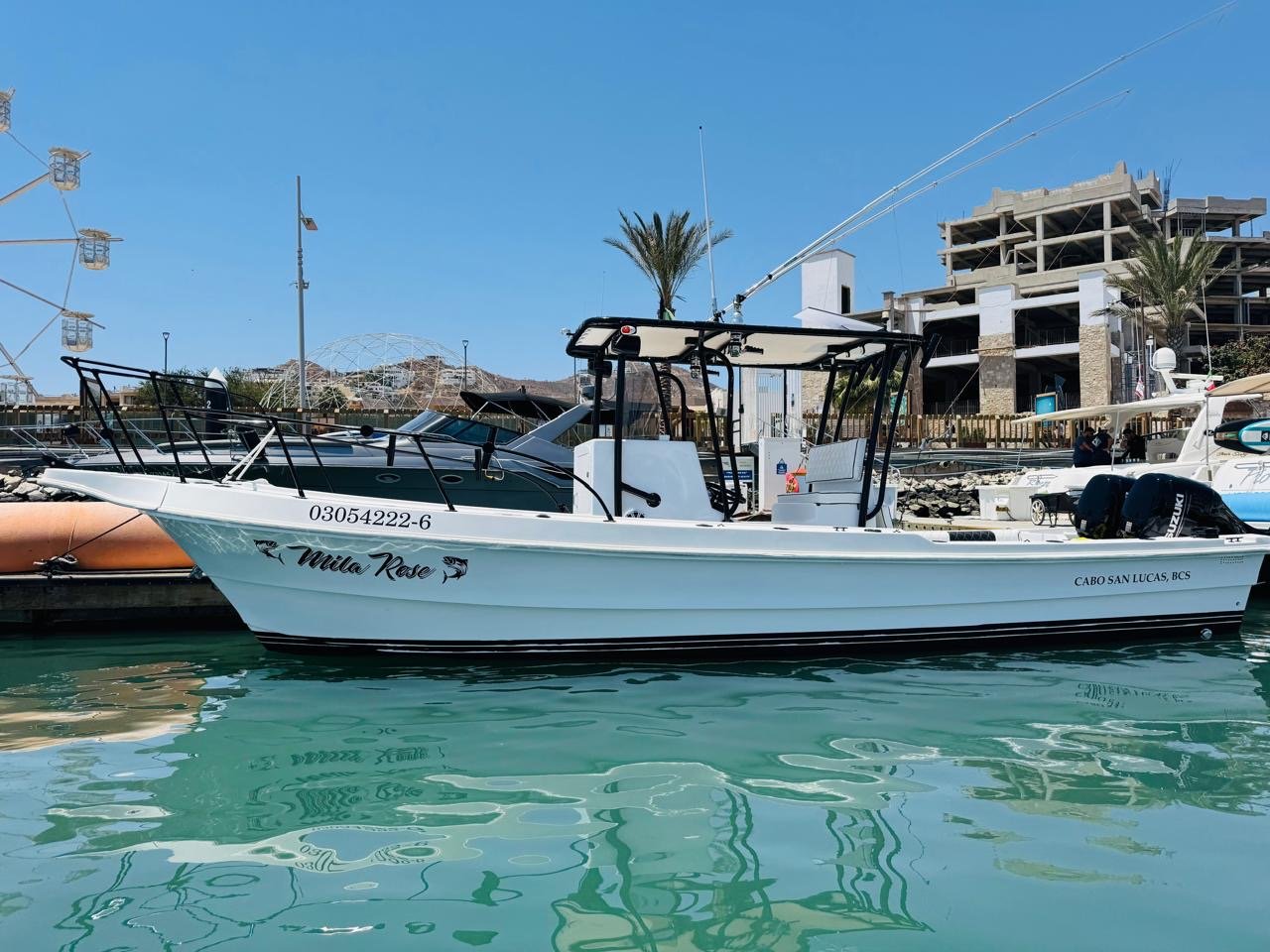 White boat named 'Mila Rose' docked at a marina with city buildings under construction in the background, and a Ferris wheel nearby, under a clear blue sky.
