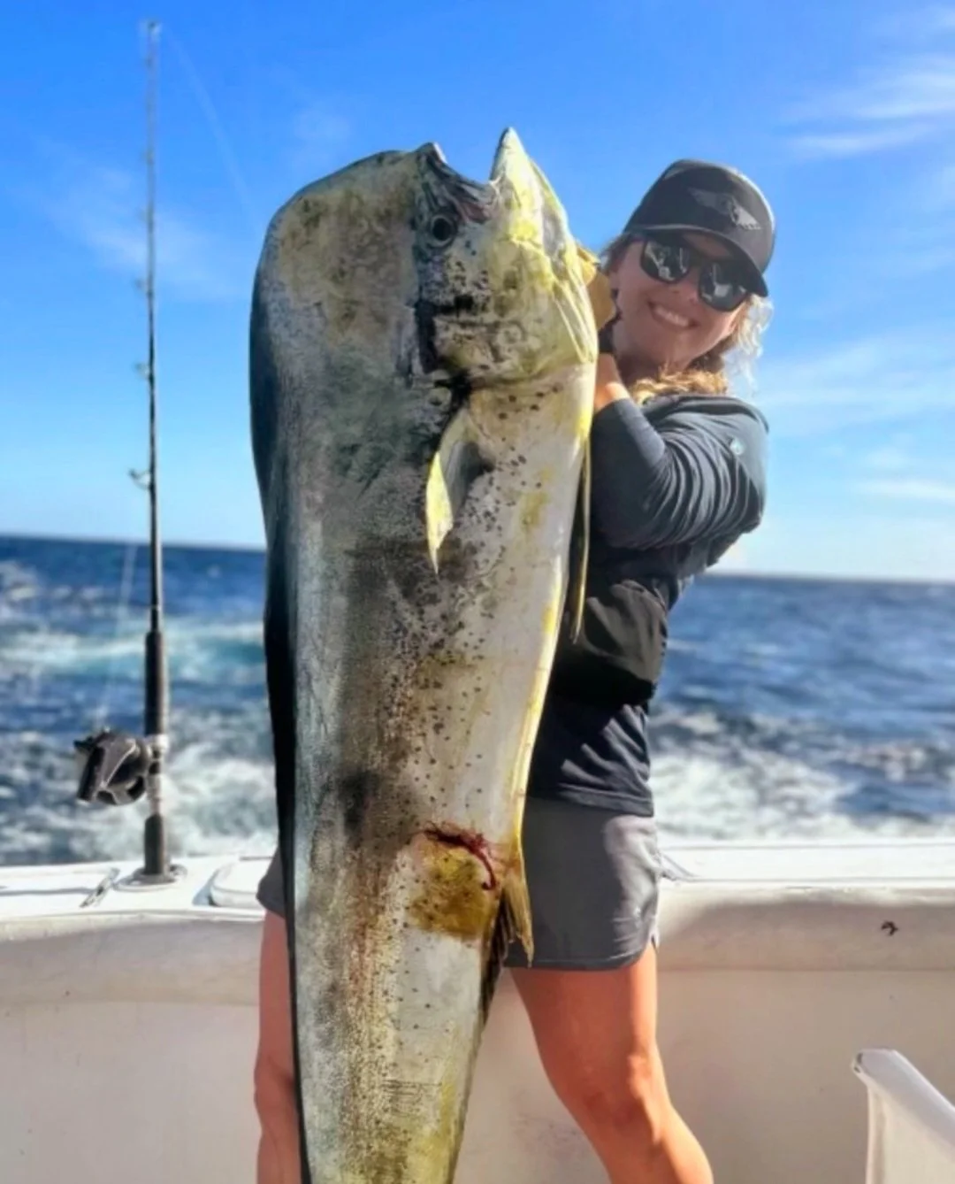 Woman on a boat holding a large fish she caught during a sunny day at sea, with fishing gear in the background.