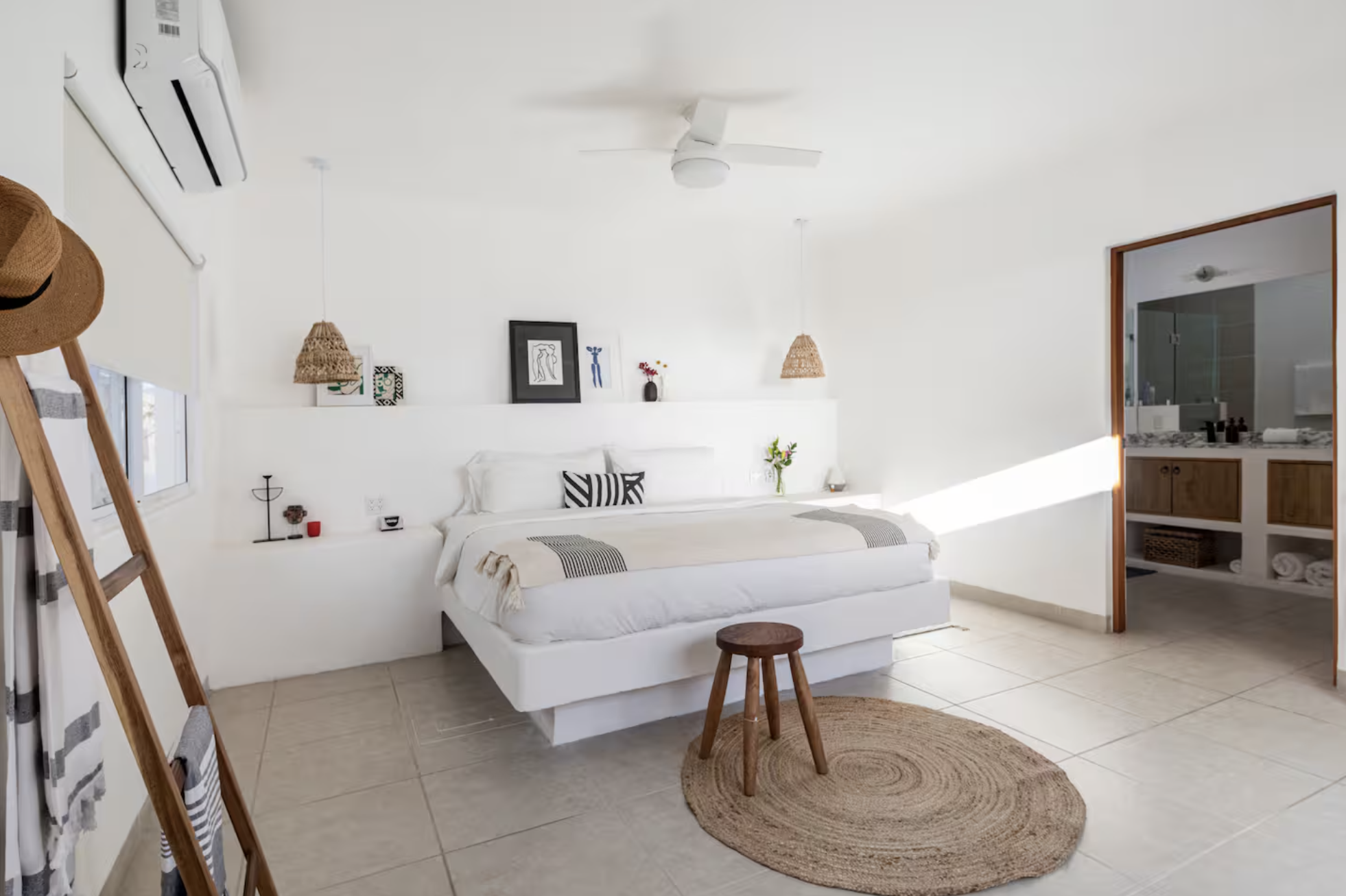 Bright hotel bedroom with white walls and minimal decor, a white bed with black and white pillows, small bedside tables with decorative items, a round woven rug, and an open door leading to a bathroom with wooden accents.