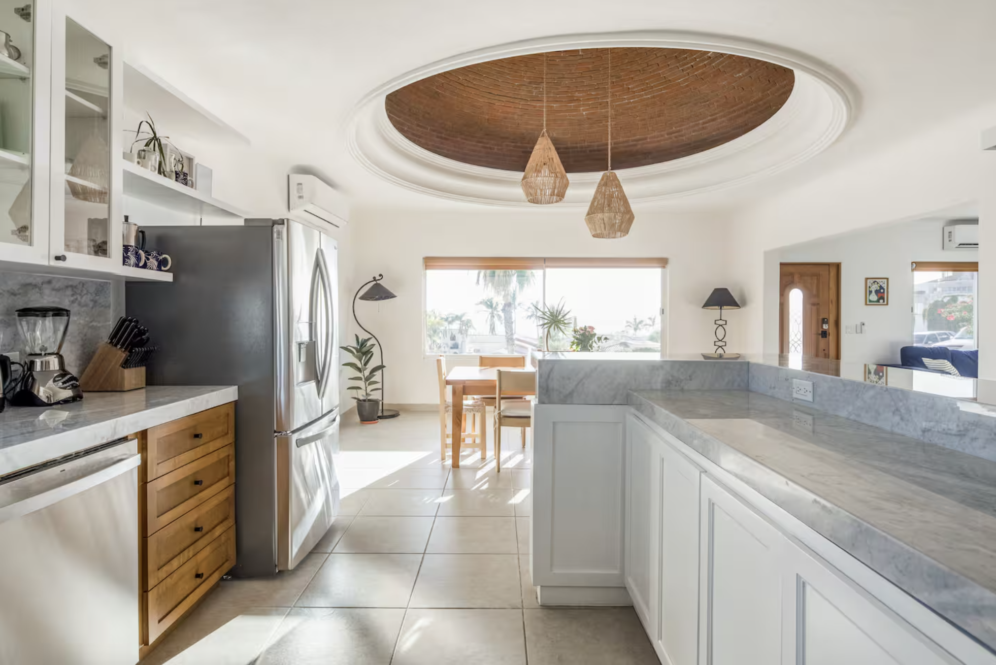Bright kitchen with white cabinets and marble countertops, dining area with wooden table and chairs, large window with a view of palm trees, ceiling with circular coffered design and hanging woven pendant lights, and a door.