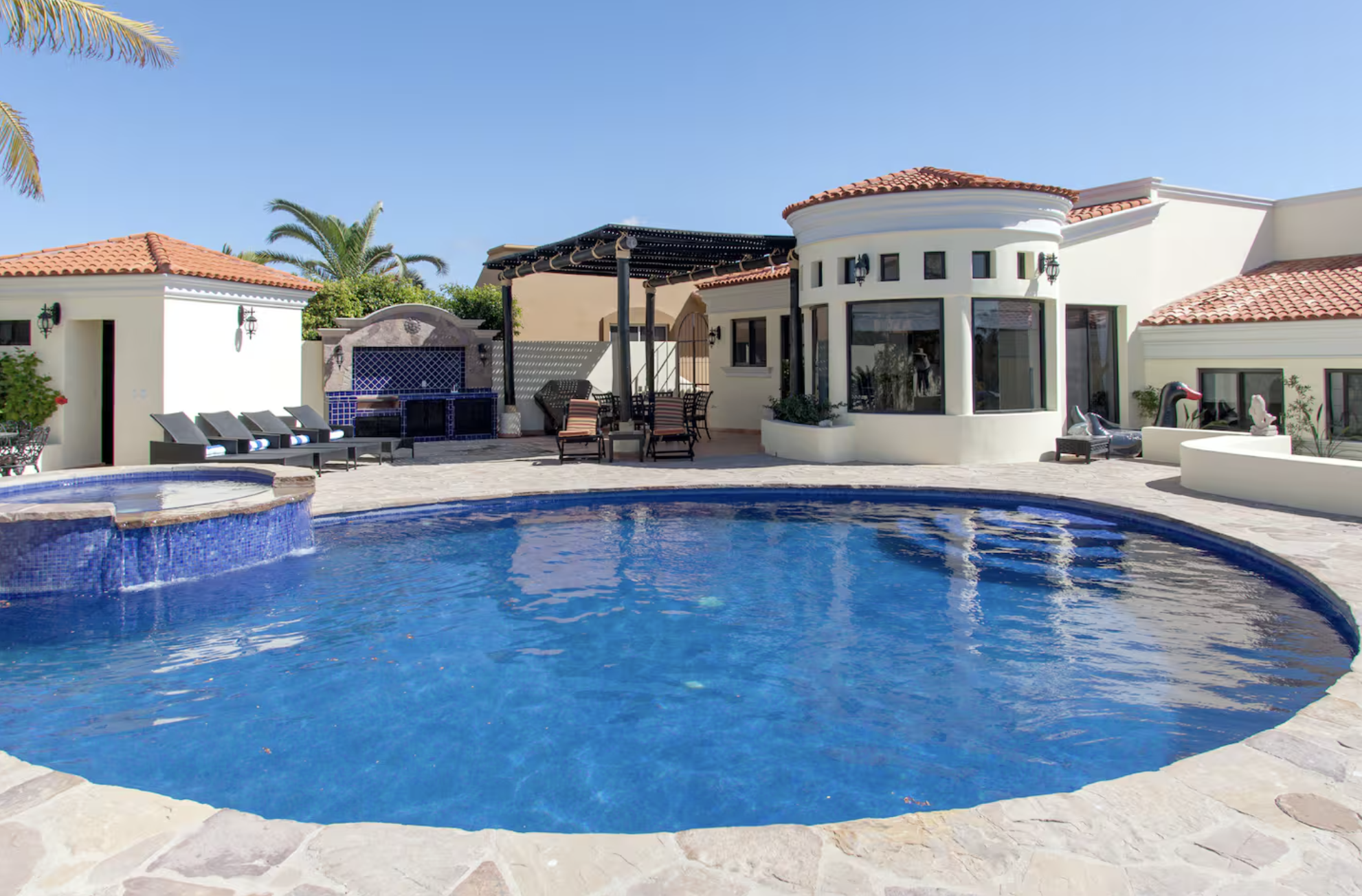 Luxury house with a curved swimming pool, multiple lounge chairs, a shaded patio area, and tropical palm trees under a clear blue sky.