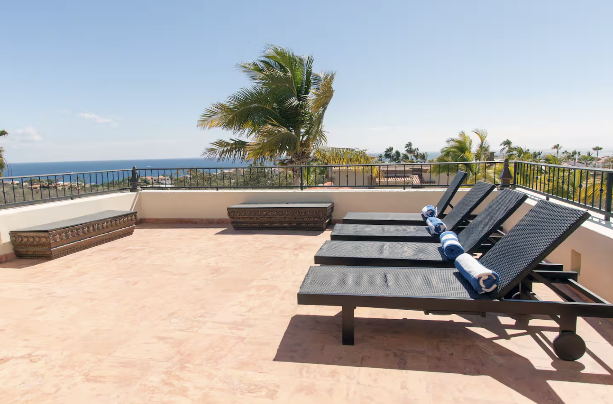 Rooftop patio with four lounge chairs, rolled towels, and decorative wooden benches, overlooking palm trees and a distant ocean on a sunny day.