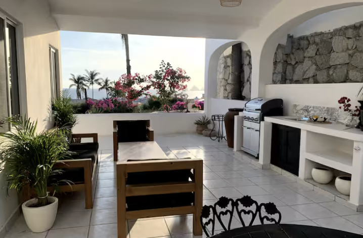 Outdoor patio area with a dining table, chairs, potted plants, and a view of palm trees and pink flowers, with a grill and kitchen counter.