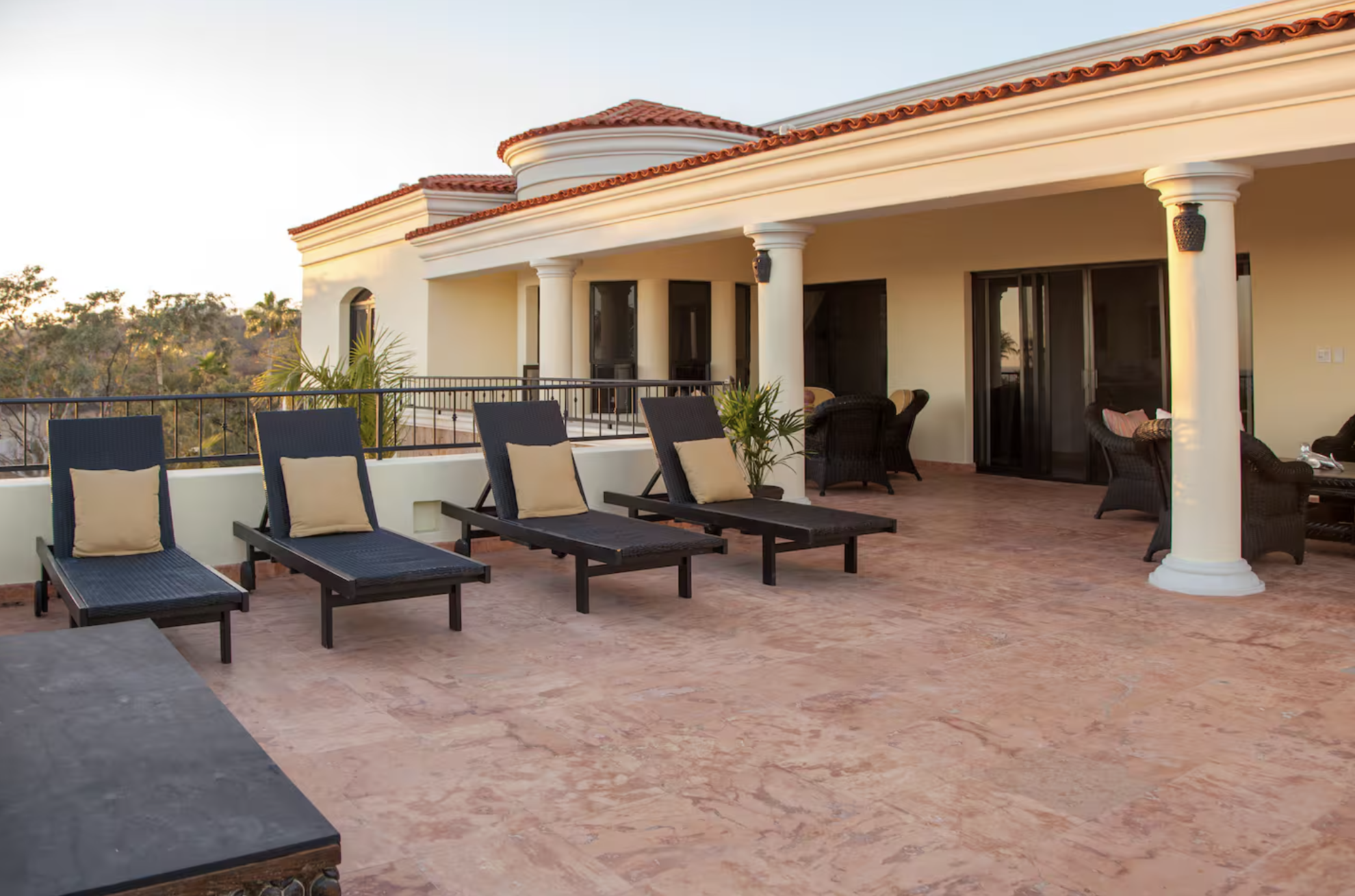 Luxury outdoor balcony with black lounge chairs and beige pillows, potted plants, and wicker seating, overlooking a tree-lined landscape at sunset.