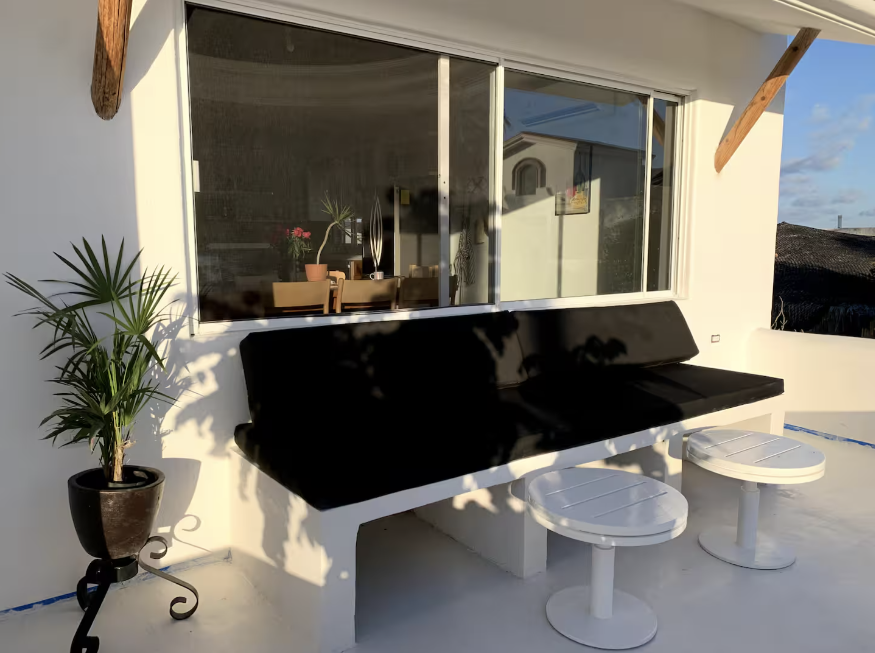 Sunlit outdoor patio with a black bench, two white round stools, potted plant, and large window reflecting neighboring buildings.