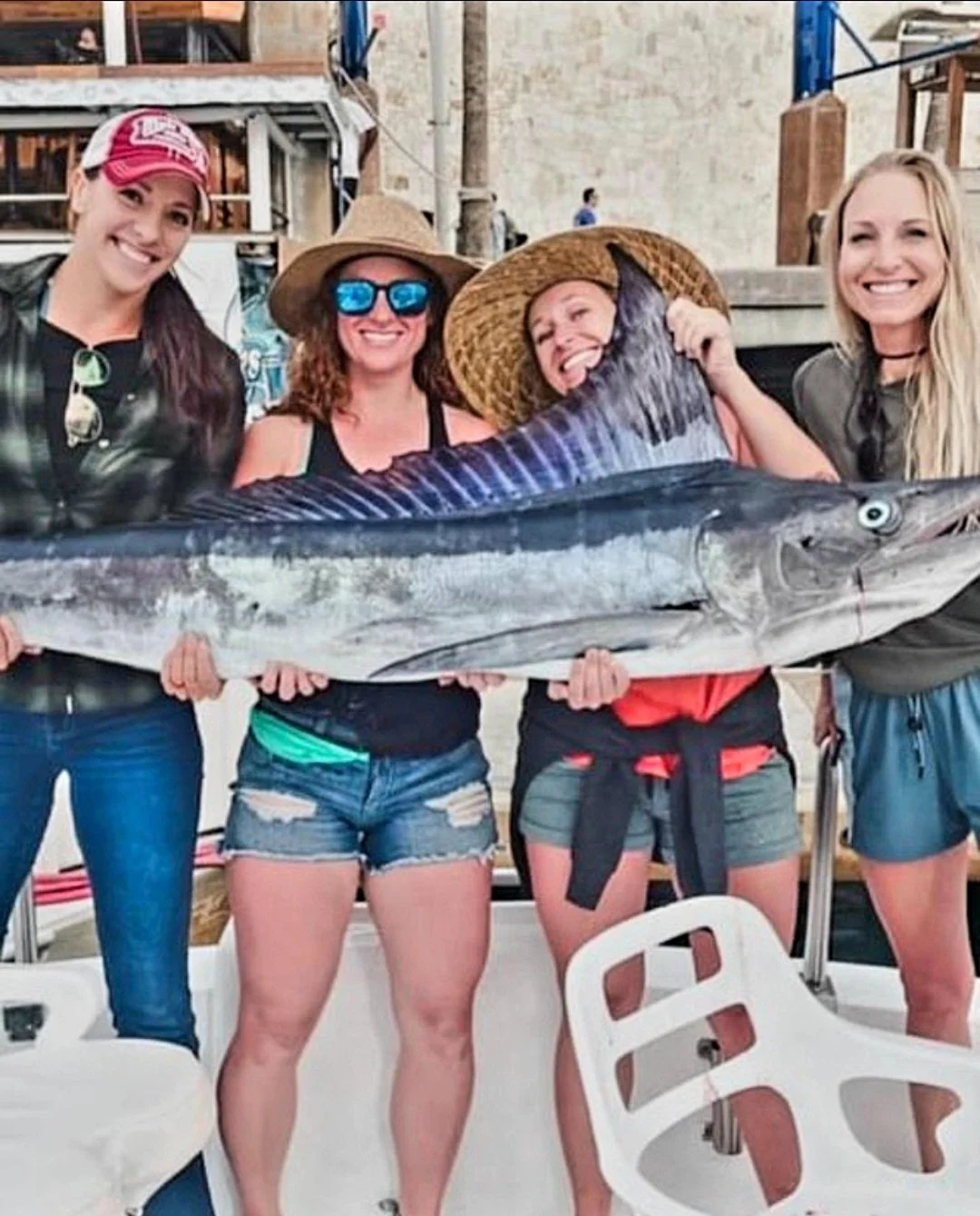 Four women holding a large fish on a boat, smiling and wearing casual summer clothes and hats. The Marlin Hunter Fishing Charter Day Trip from Cabo