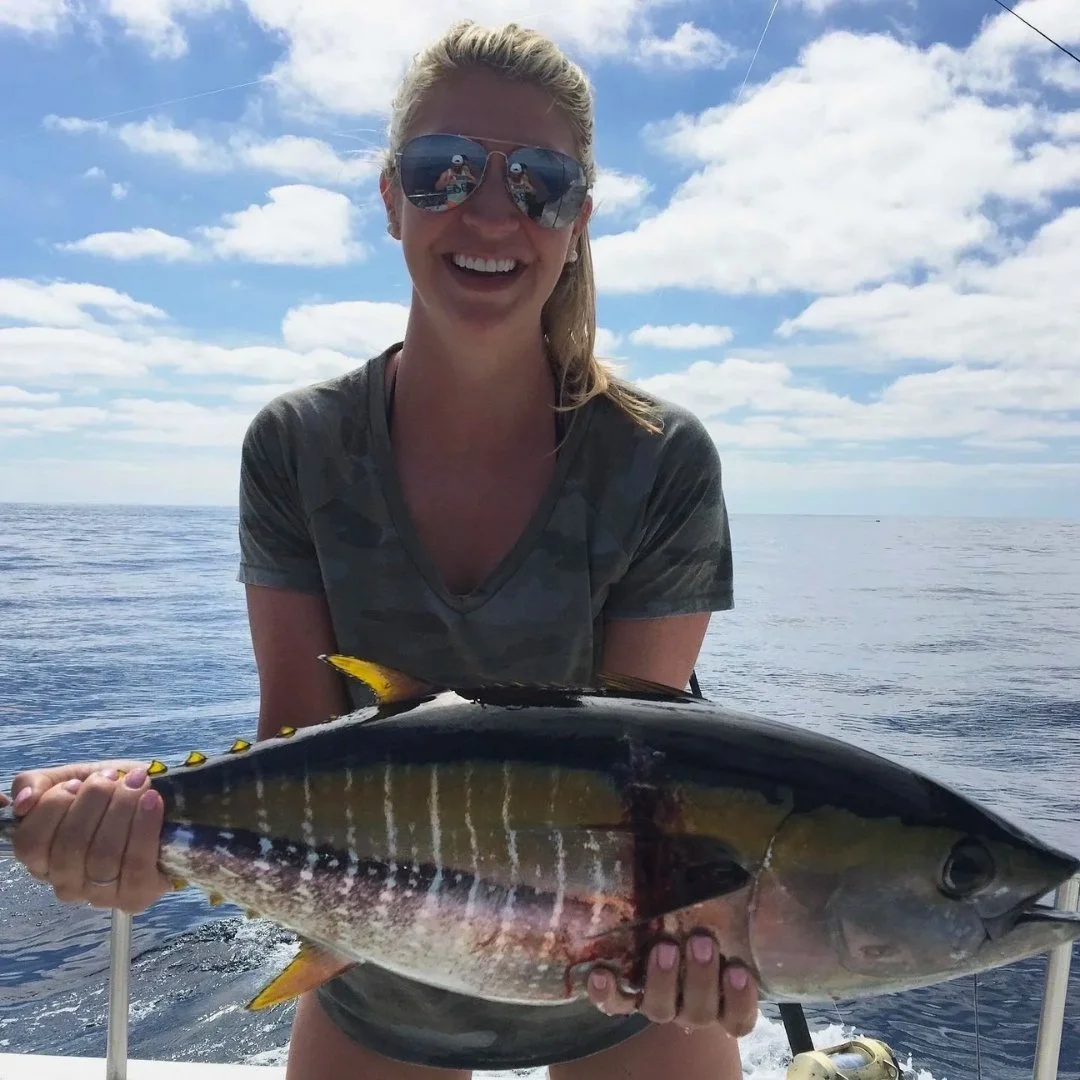 A woman on a boat holding a large fish she caught, with a smiling face in sunglasses, blue sky, and ocean in background. The Marlin Hunter Fishing Charter 
